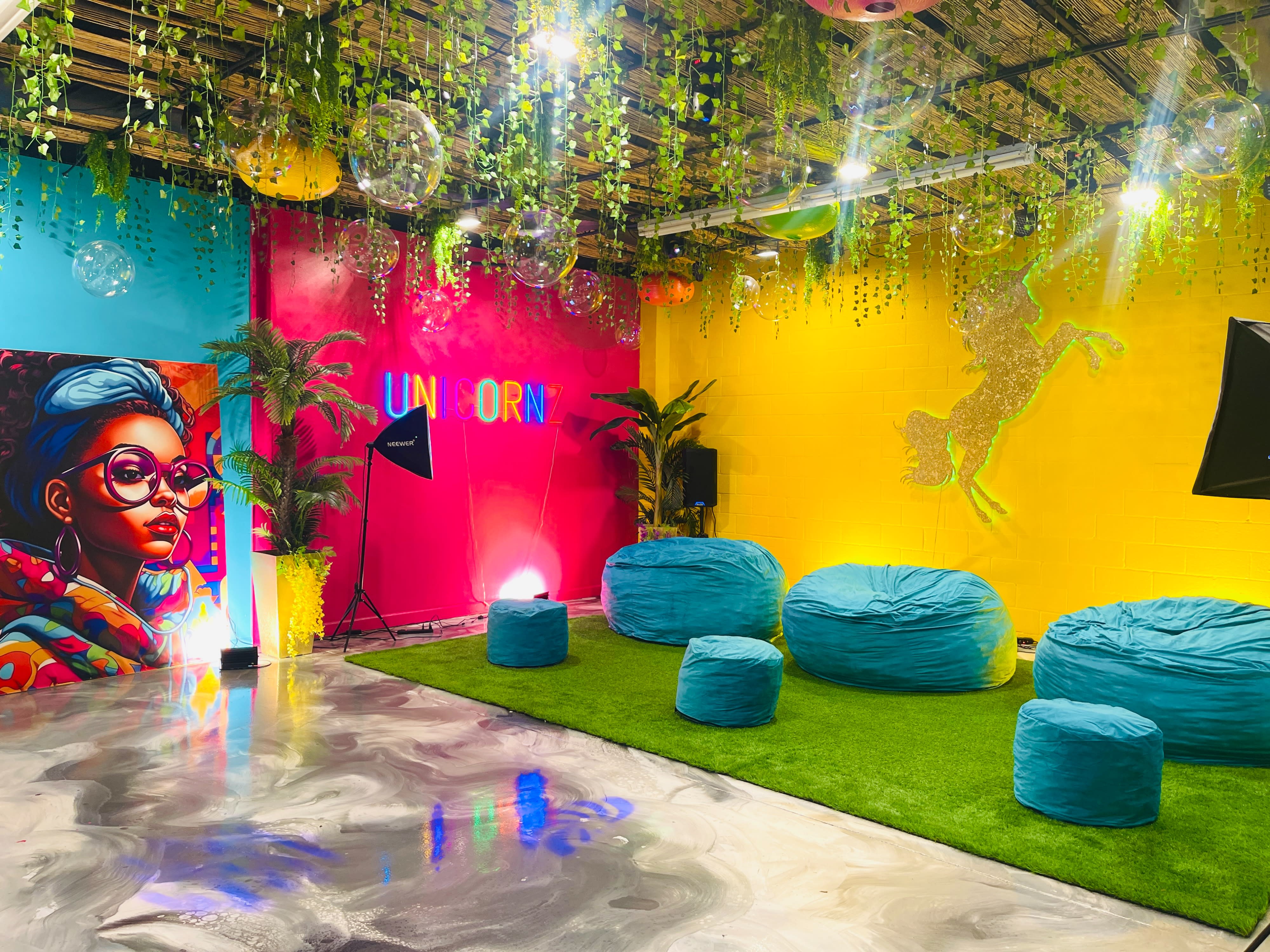 A woman and a girl pose together on cozy chairs in a colorful photo shoot setting adorned with bright decorations and greenery.