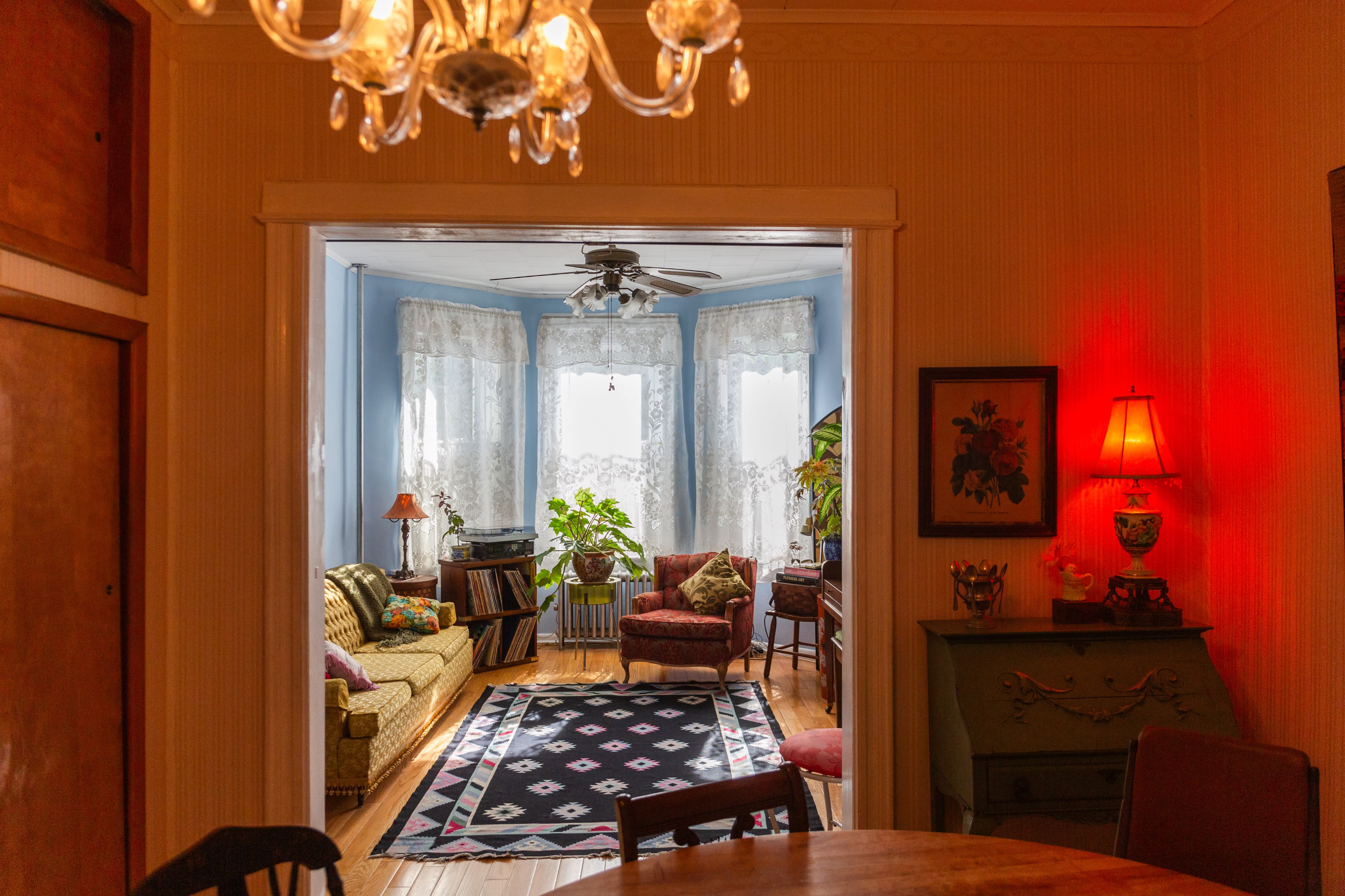 An editorial photo shoot captures a vintage sitting area featuring a tufted pink sofa, a decorative floral lamp, and a mirror reflecting the ornate wallpaper and artwork.