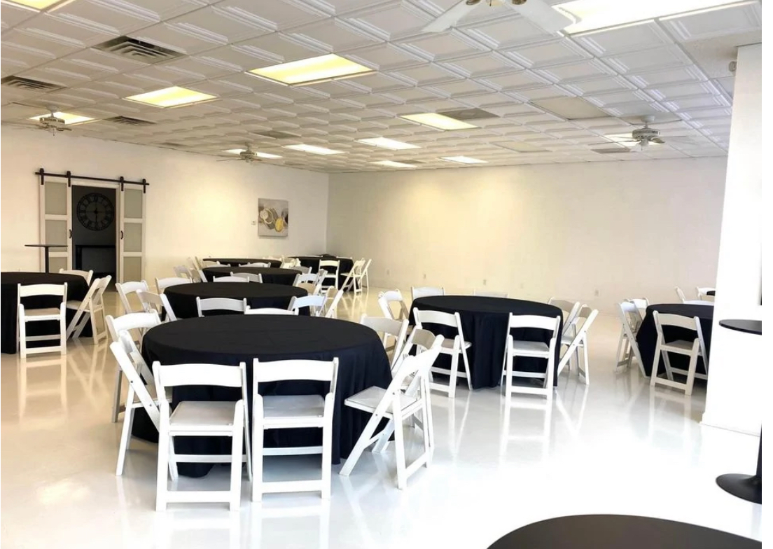 The spacious venue is set up for a baby shower, featuring decorated tables with pink and white accents and a backdrop adorned with balloons.