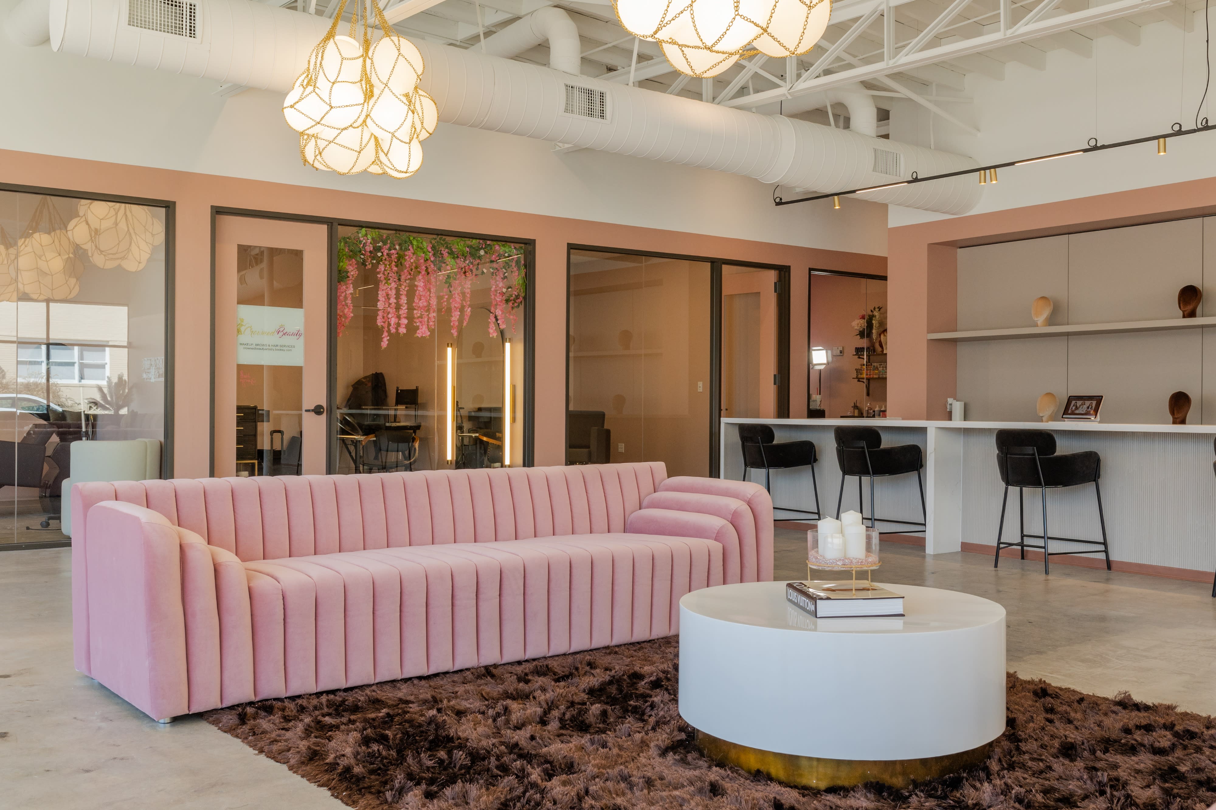 A bridal shower is taking place in a modern venue with guests sitting on a pink couch and others gathered around a table preparing food and decorations.