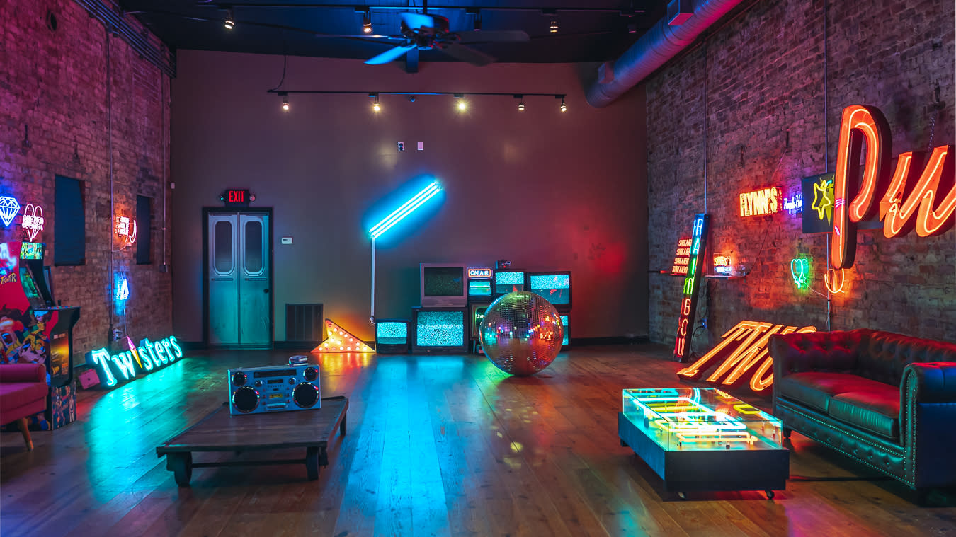 Three men sit on a couch during a video shoot, surrounded by gaming consoles and neon lights.