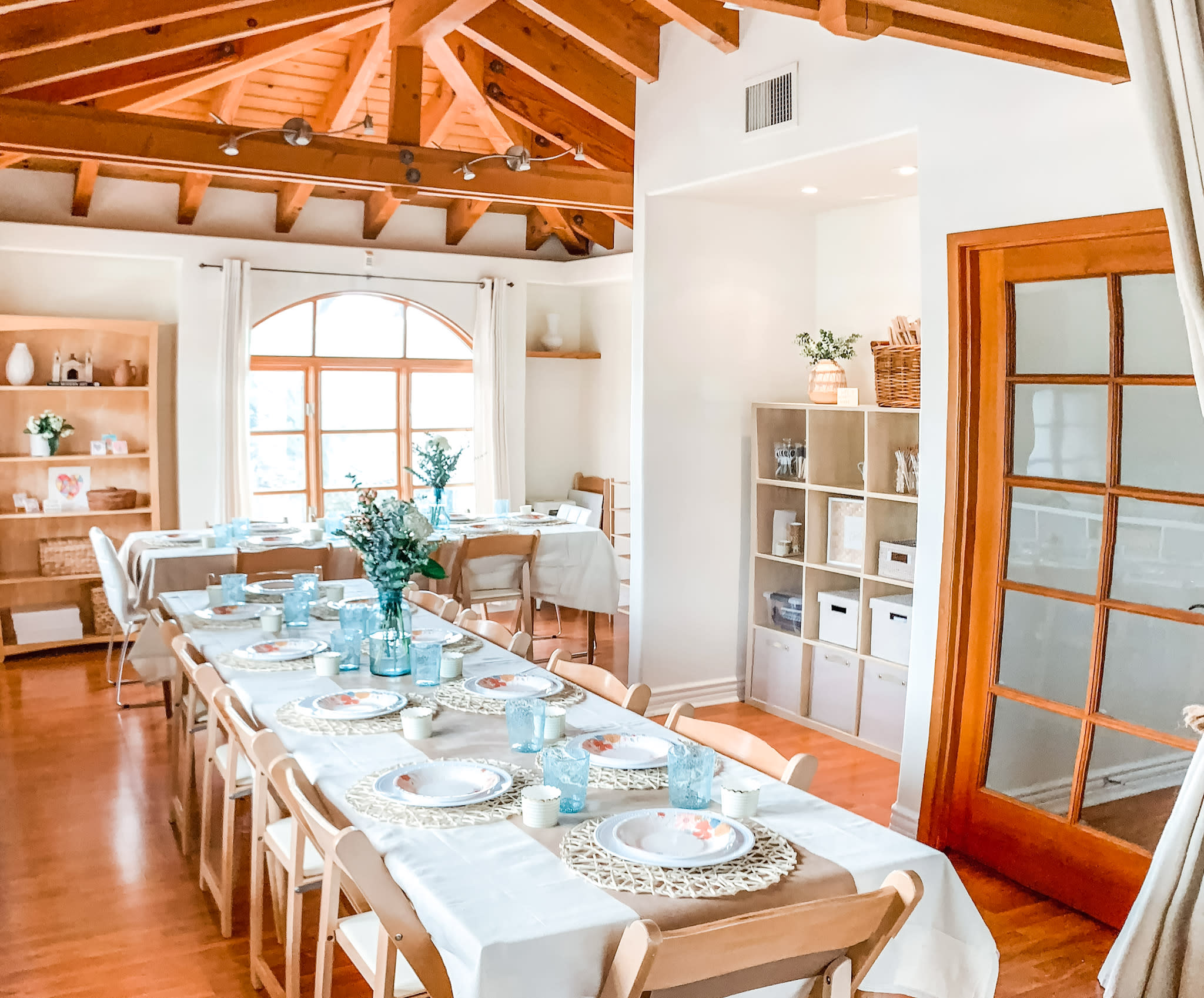 A long table is set in a room adorned with flowers and framed photos, overlooking a scenic view through a large arched window.
