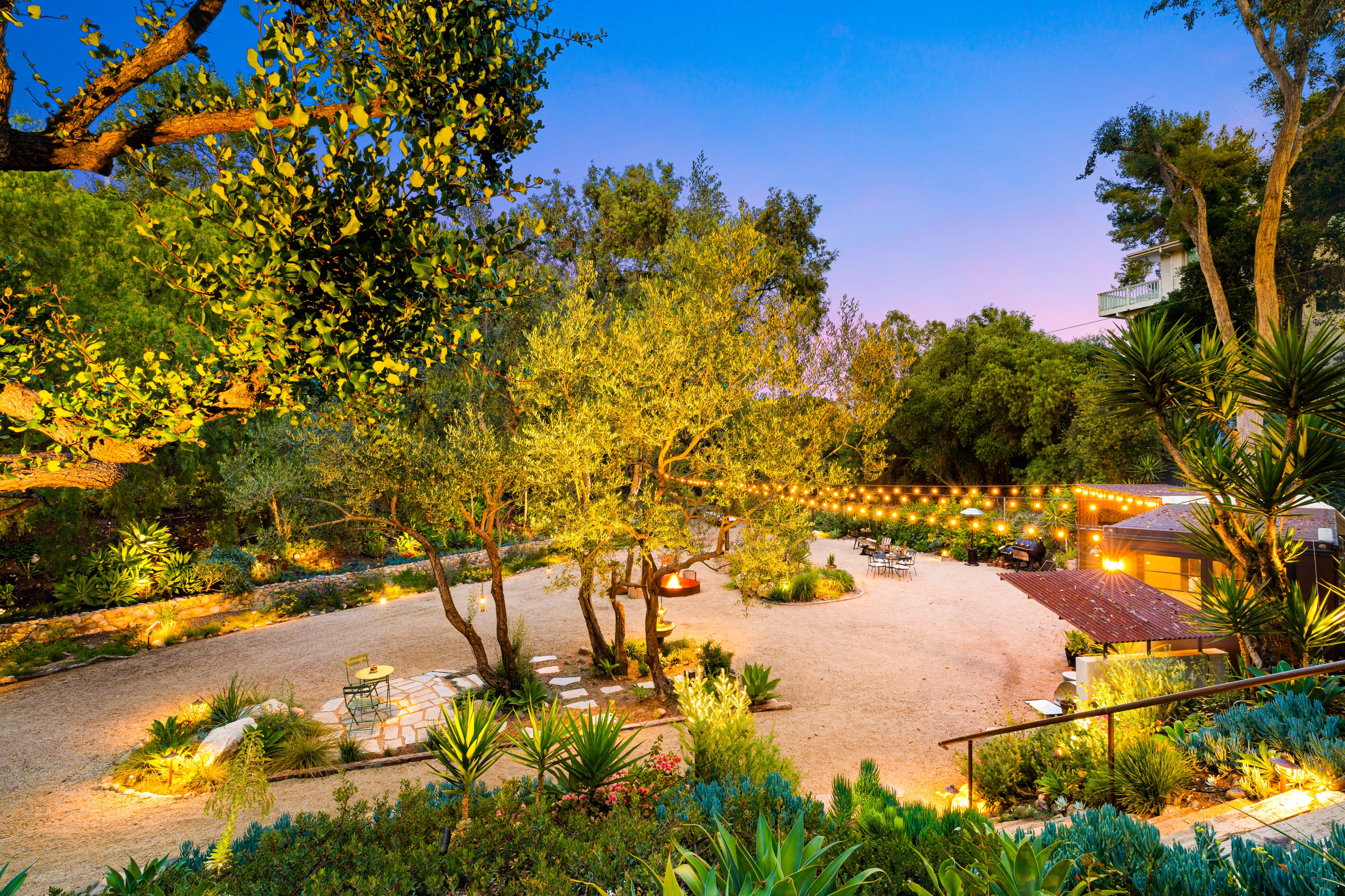 A group of people gathers around a fire pit in a landscaped outdoor setting, enjoying drinks and conversation during an engagement party.