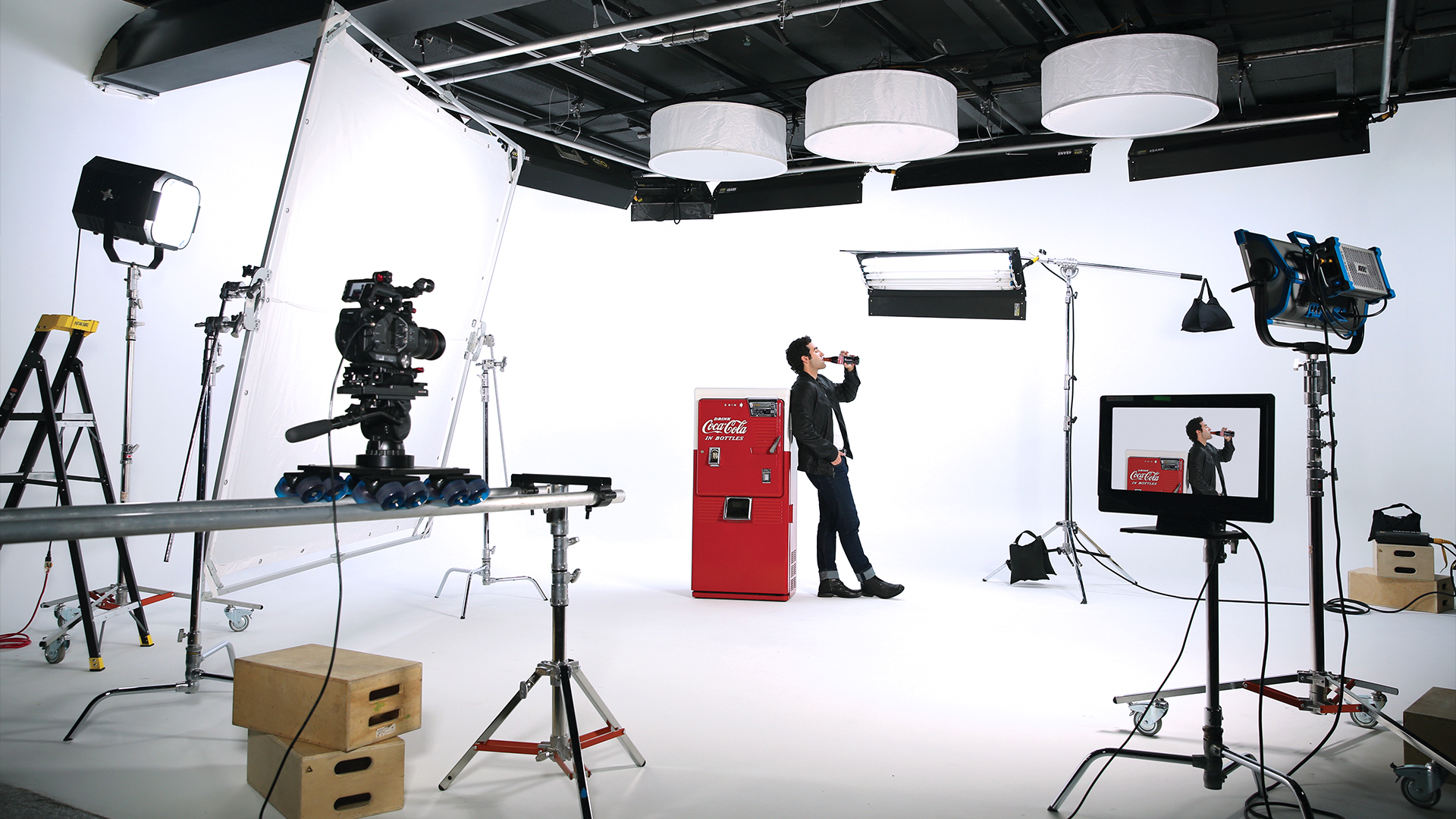 A person sits in a director's chair while a camera is positioned on a tripod in a well-lit video studio with a blank white backdrop.
