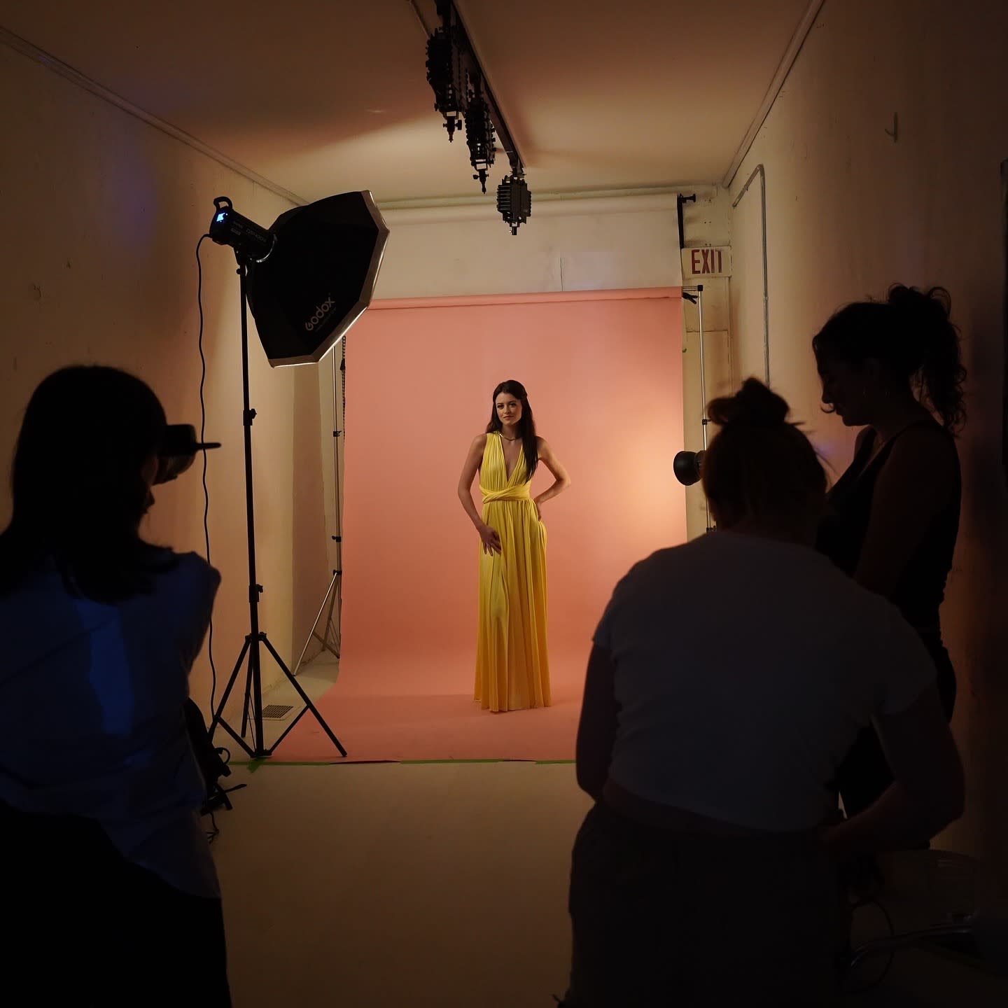A person poses seated on a chair against a solid pastel background during a photoshoot.