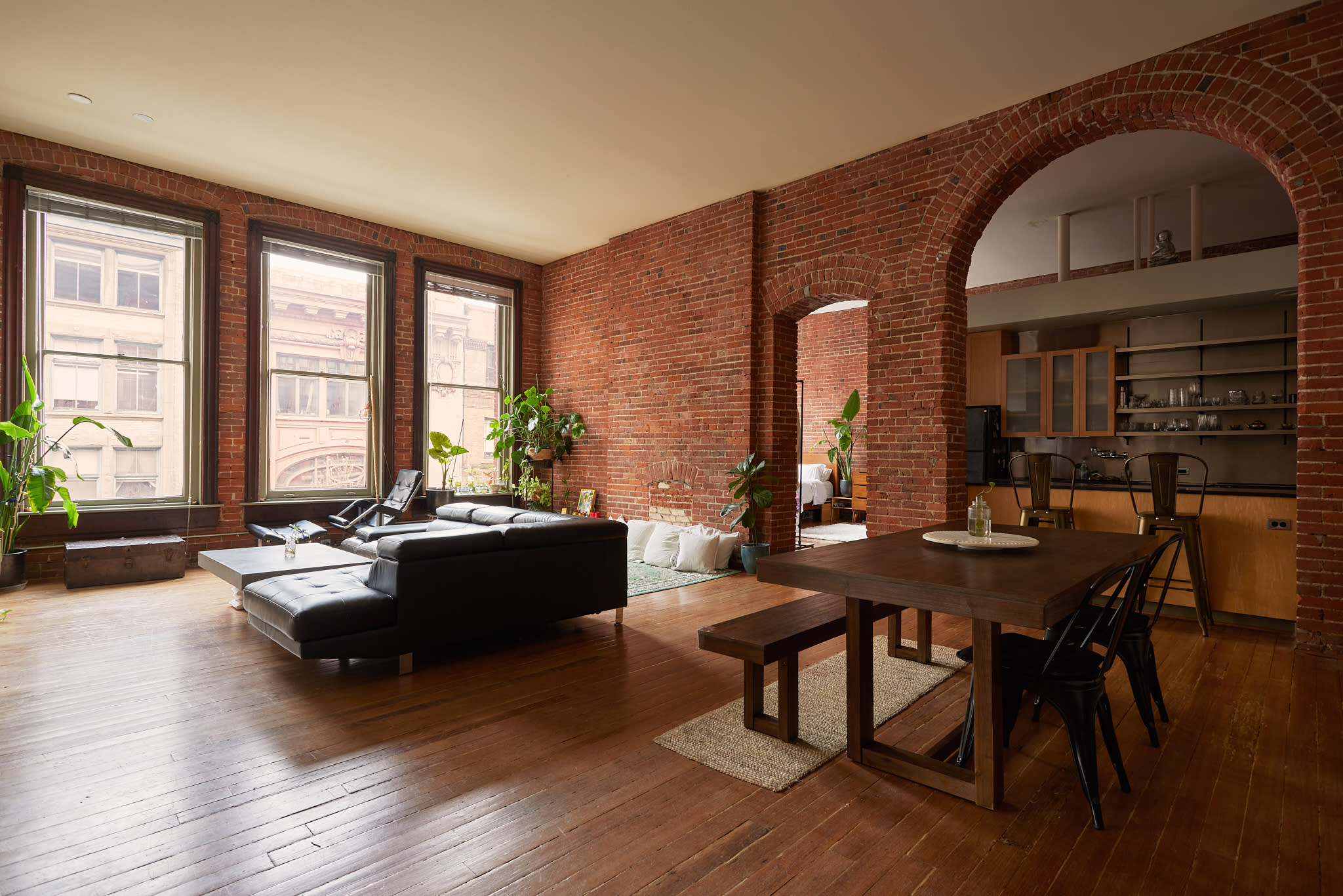 A black leather sectional sofa sits in a sunlit room with exposed brick walls, featuring large windows that overlook a cityscape.