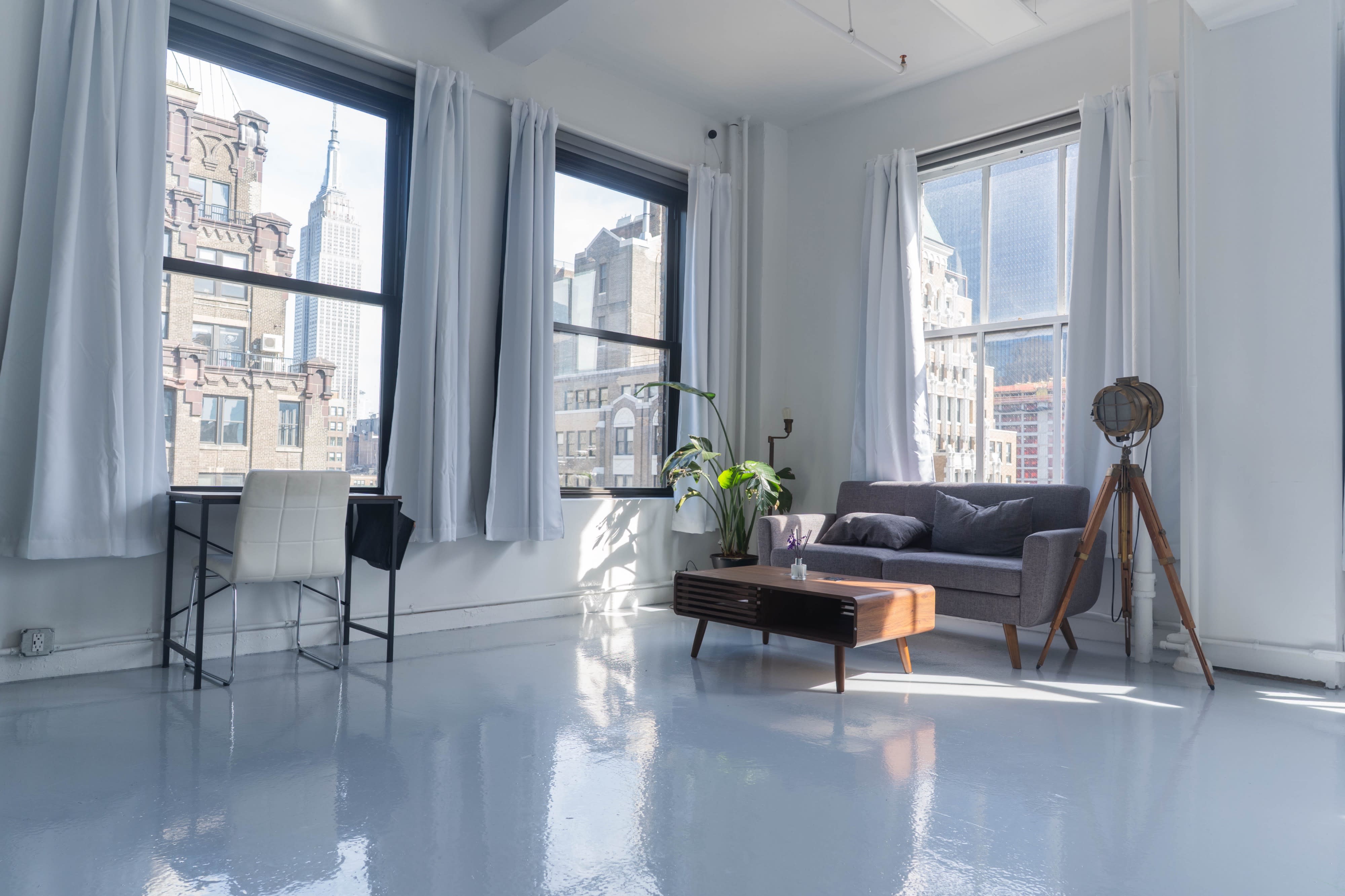 A cozy living space features a gray sofa, a wooden coffee table with a laptop, and large windows showcasing a city skyline.
