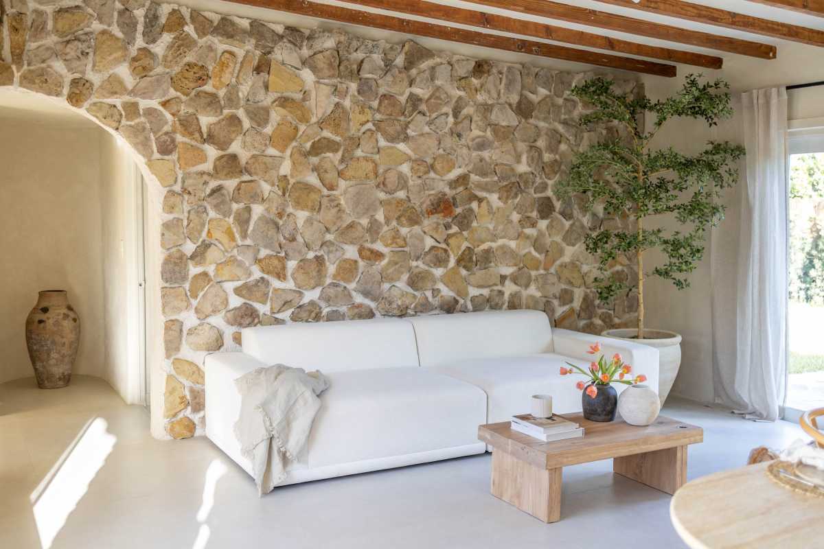 A woman sits on a white sofa in a stone-walled room, reading a book while a potted plant sits nearby.