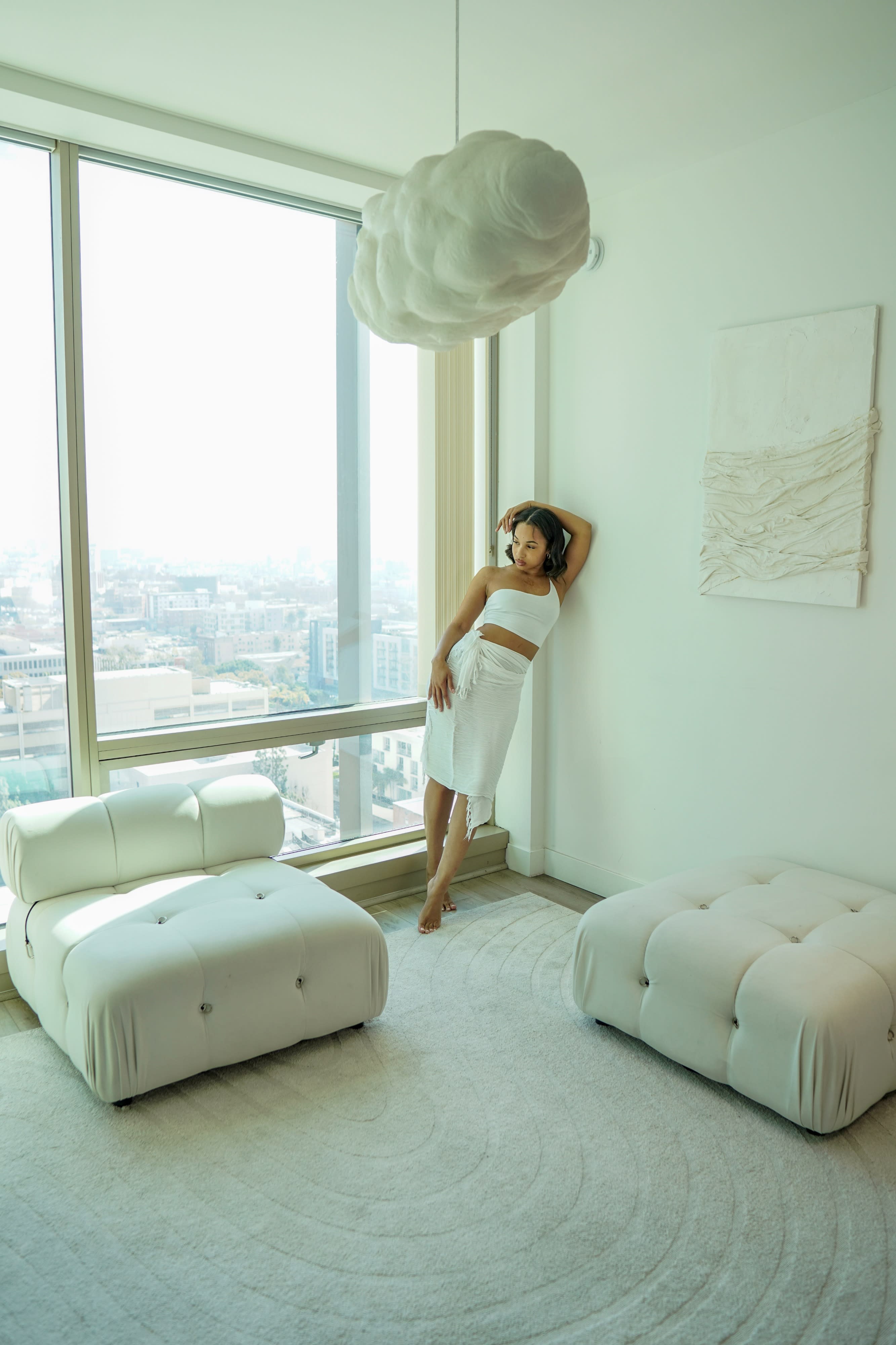 A model poses in a black off-shoulder dress while seated on a white couch in a minimalist setting.