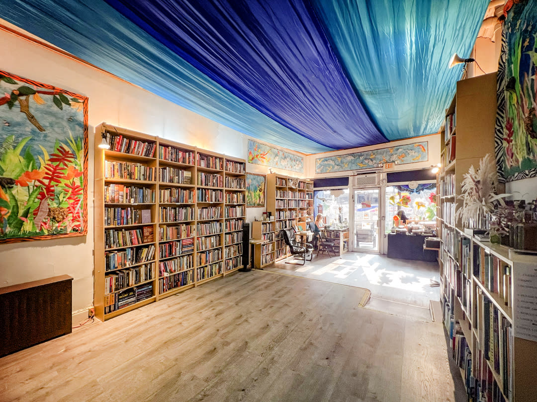 A woman in a light-colored dress reads poetry to an audience seated in a bookstore filled with books.