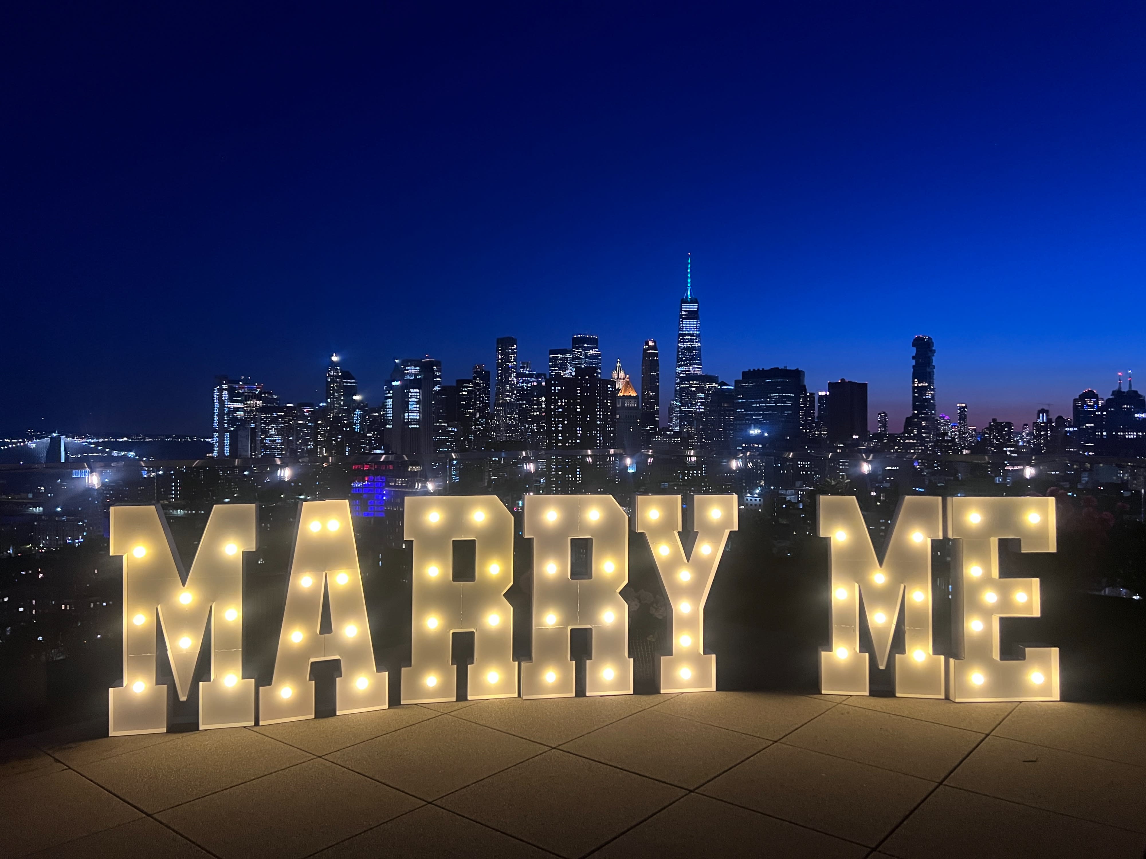 A man kneels in front of a woman, proposing with a ring on a rooftop adorned with flowers, overlooking a city skyline at sunset.
