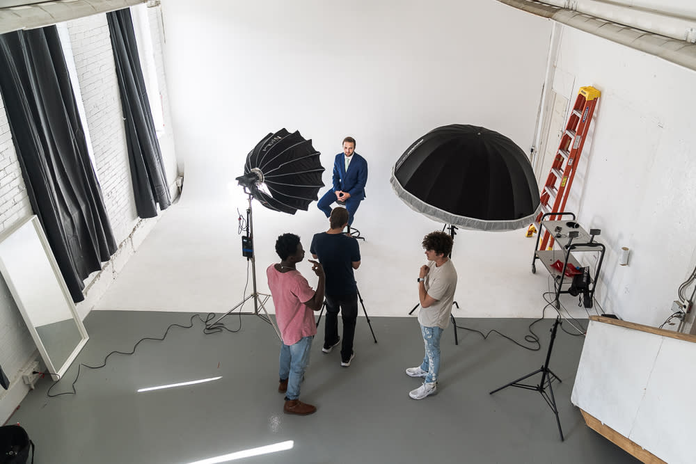 A woman poses thoughtfully while seated on a director's chair during a photo shoot.