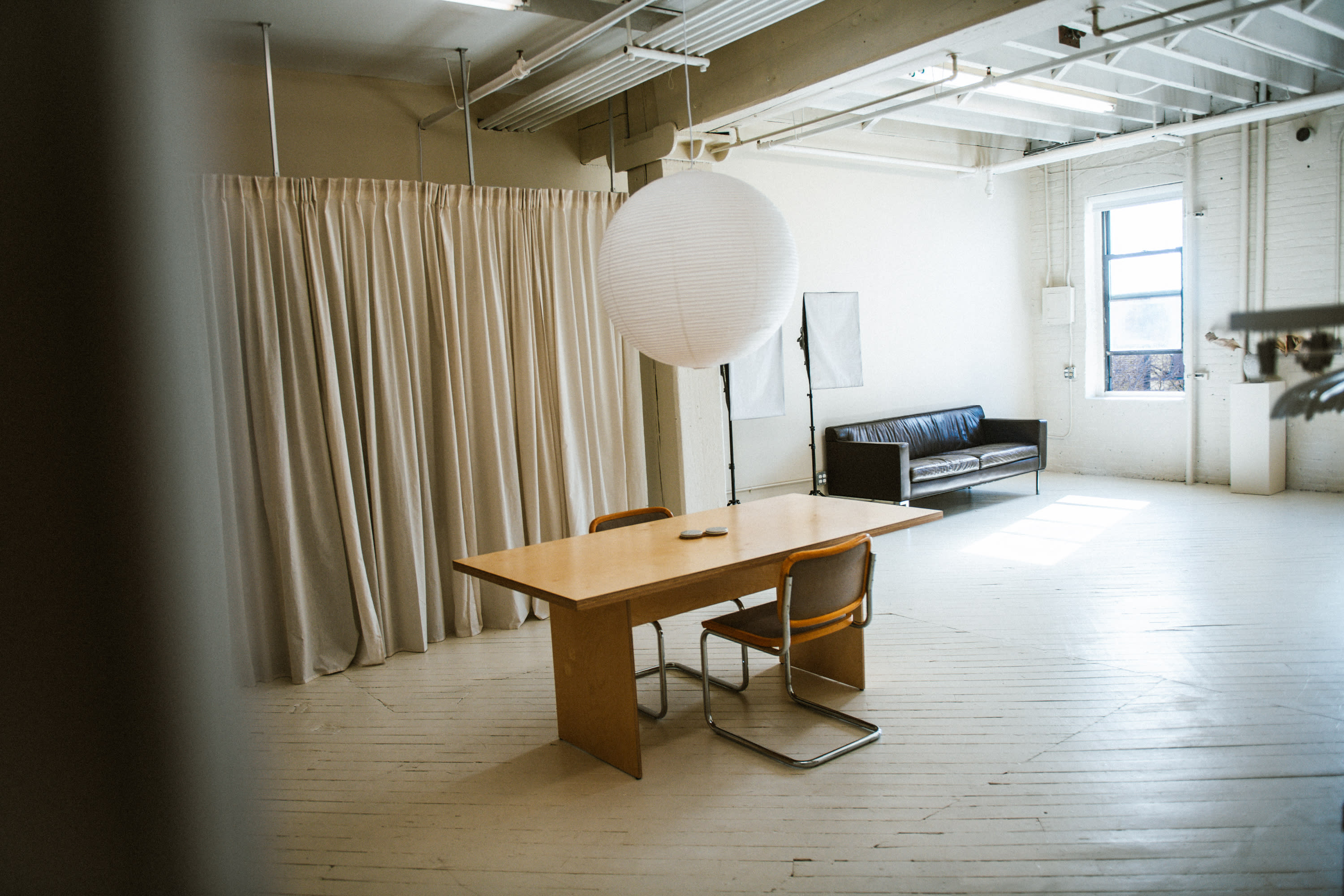 A minimalist workspace features a wooden table with a laptop and notebooks, illuminated by a hanging paper lantern.