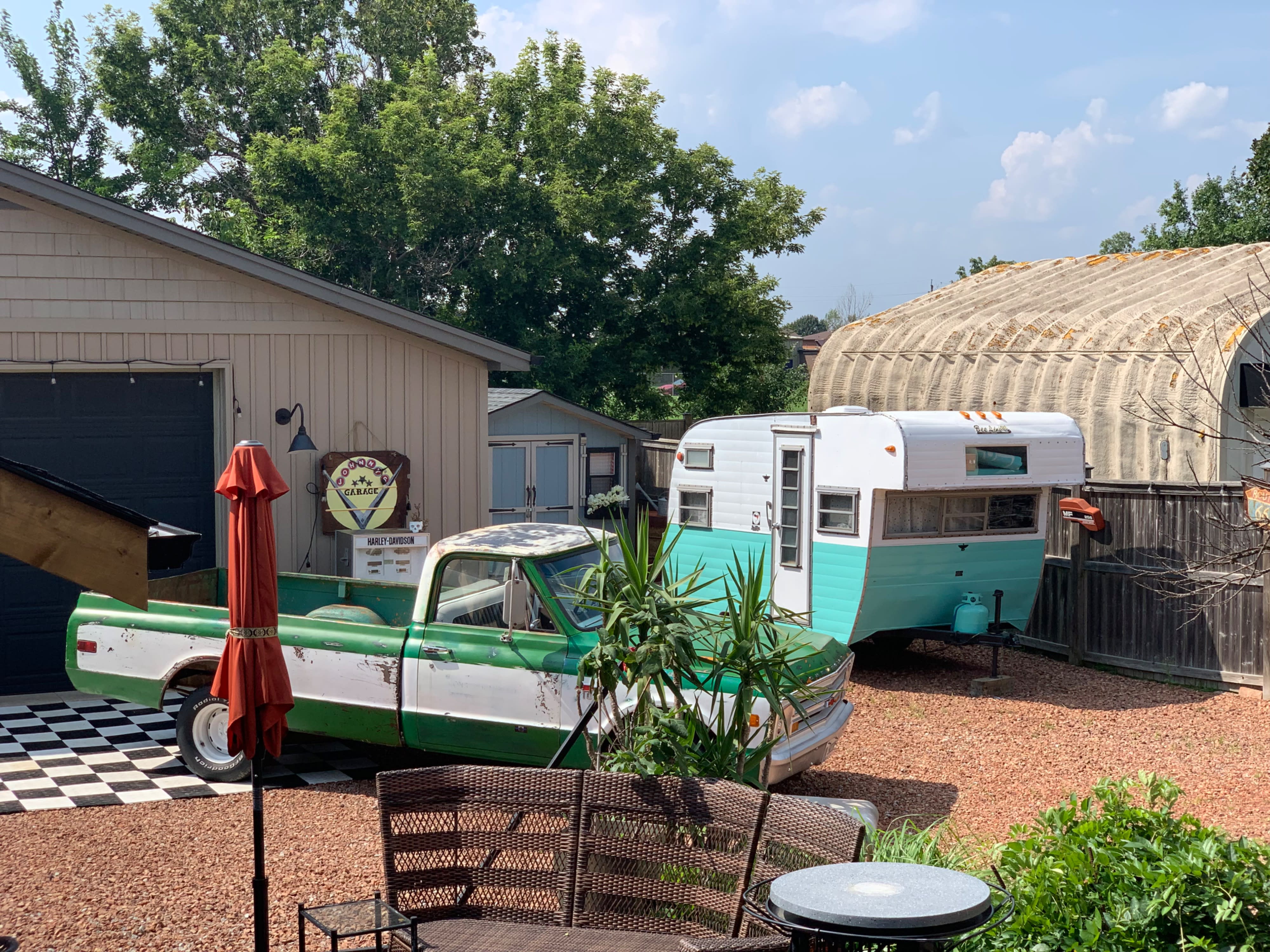 A photographer kneels on the ground as six models pose in front of a vintage trailer during an outdoor video shoot.