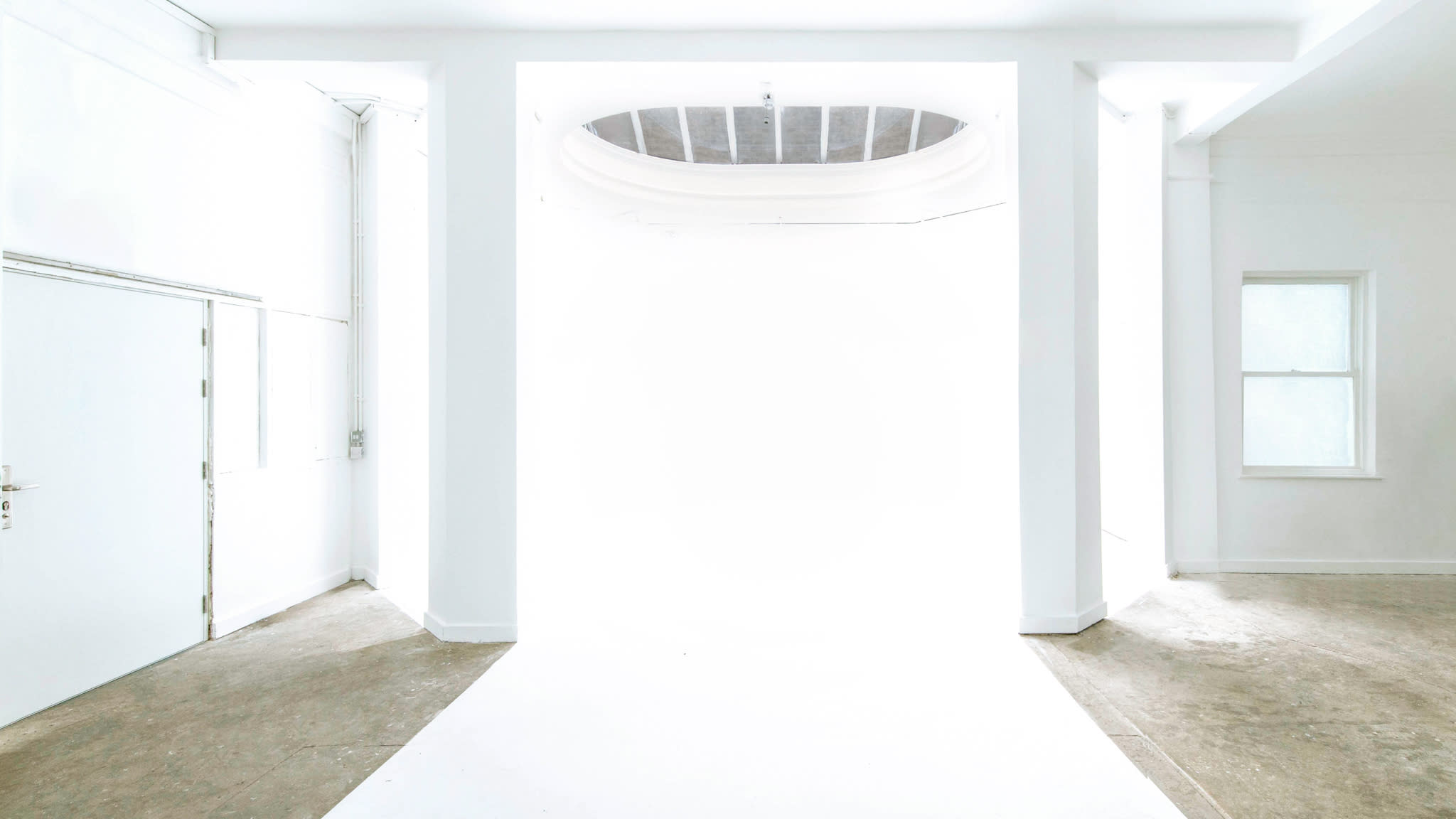 A person stands behind a wall while another individual adjusts a camera on a tripod in a minimalistic studio space.