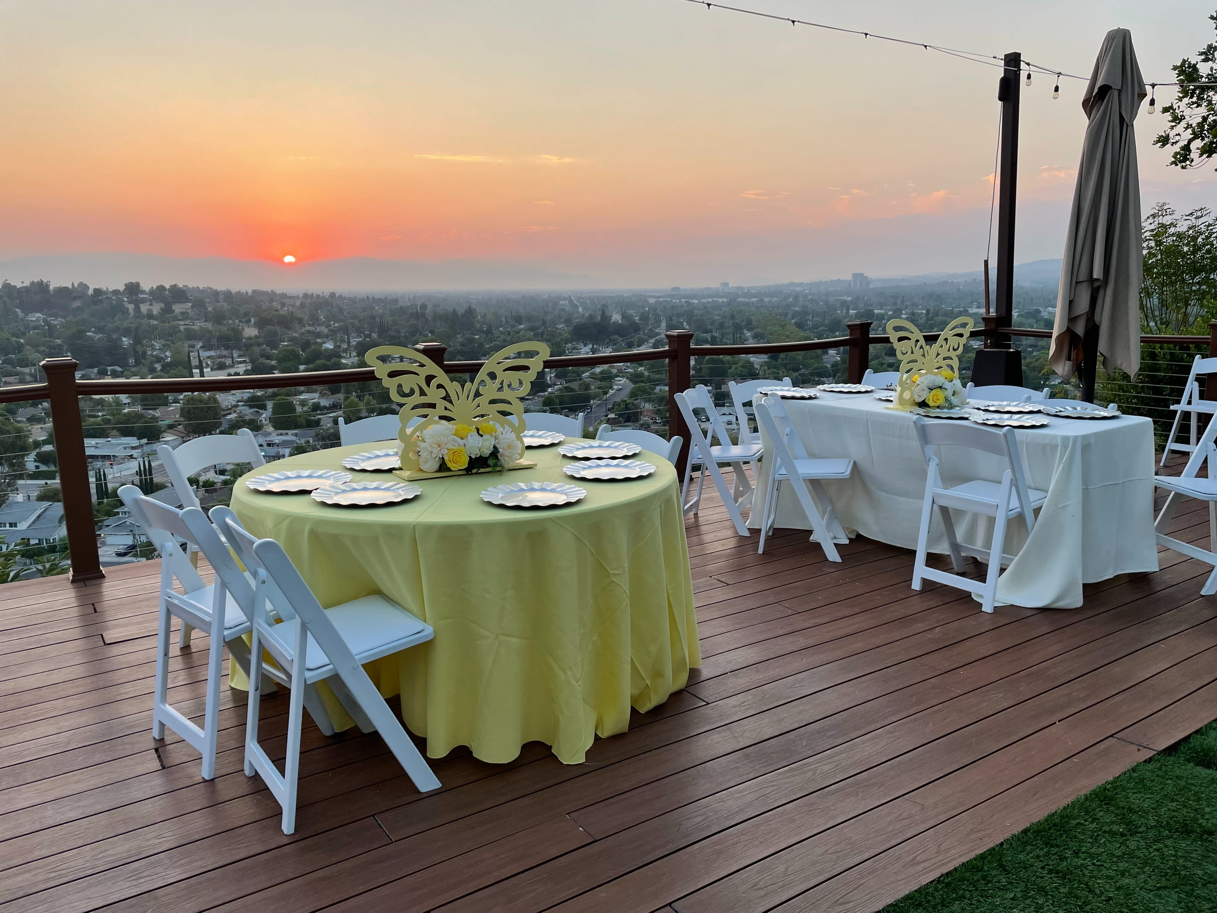 A pregnant woman in a pink dress stands beside a man seated in a folding chair during a baby shower celebration on a patio with a scenic view.