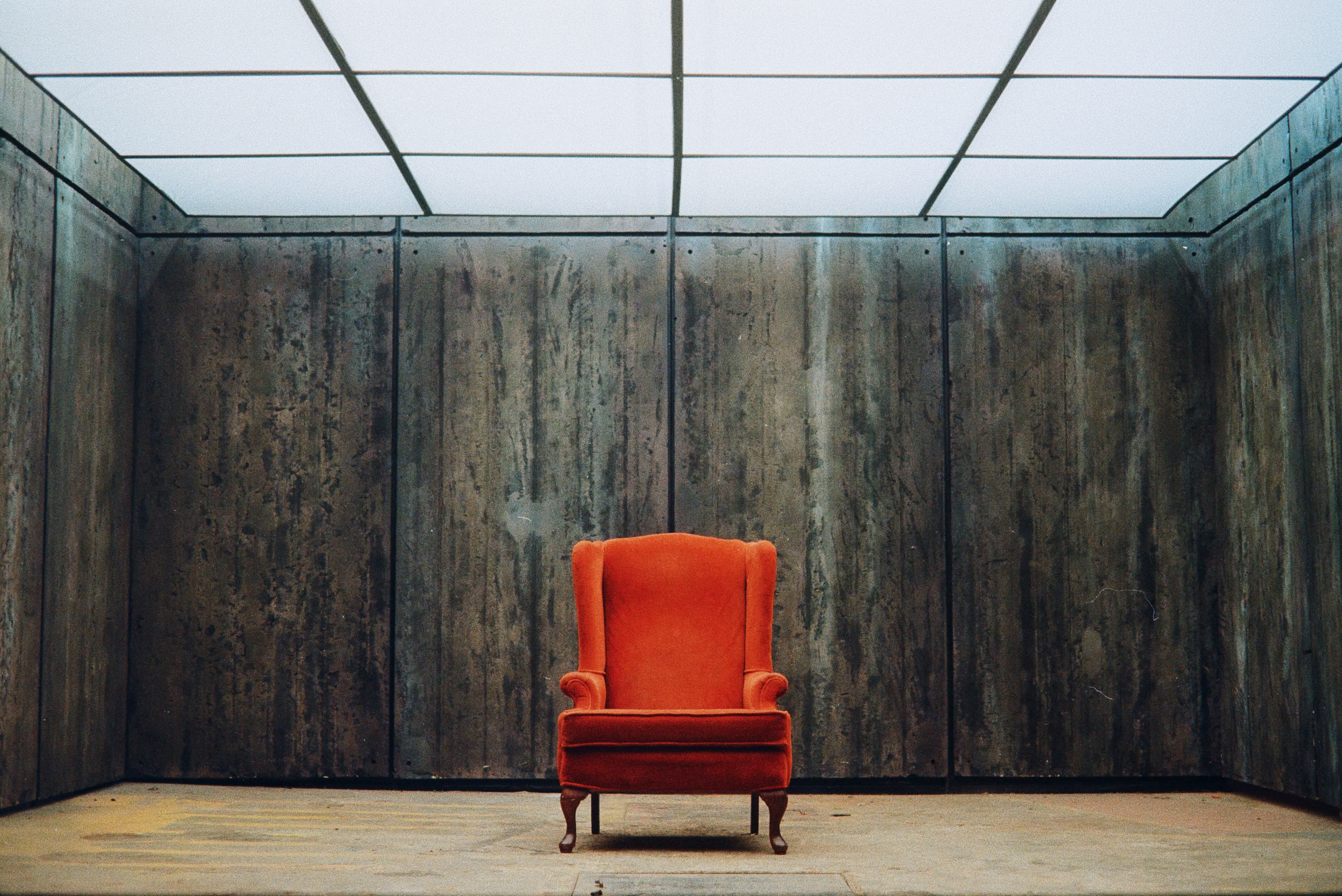 An orange armchair is placed in the center of a dimly lit, industrial room with concrete walls.