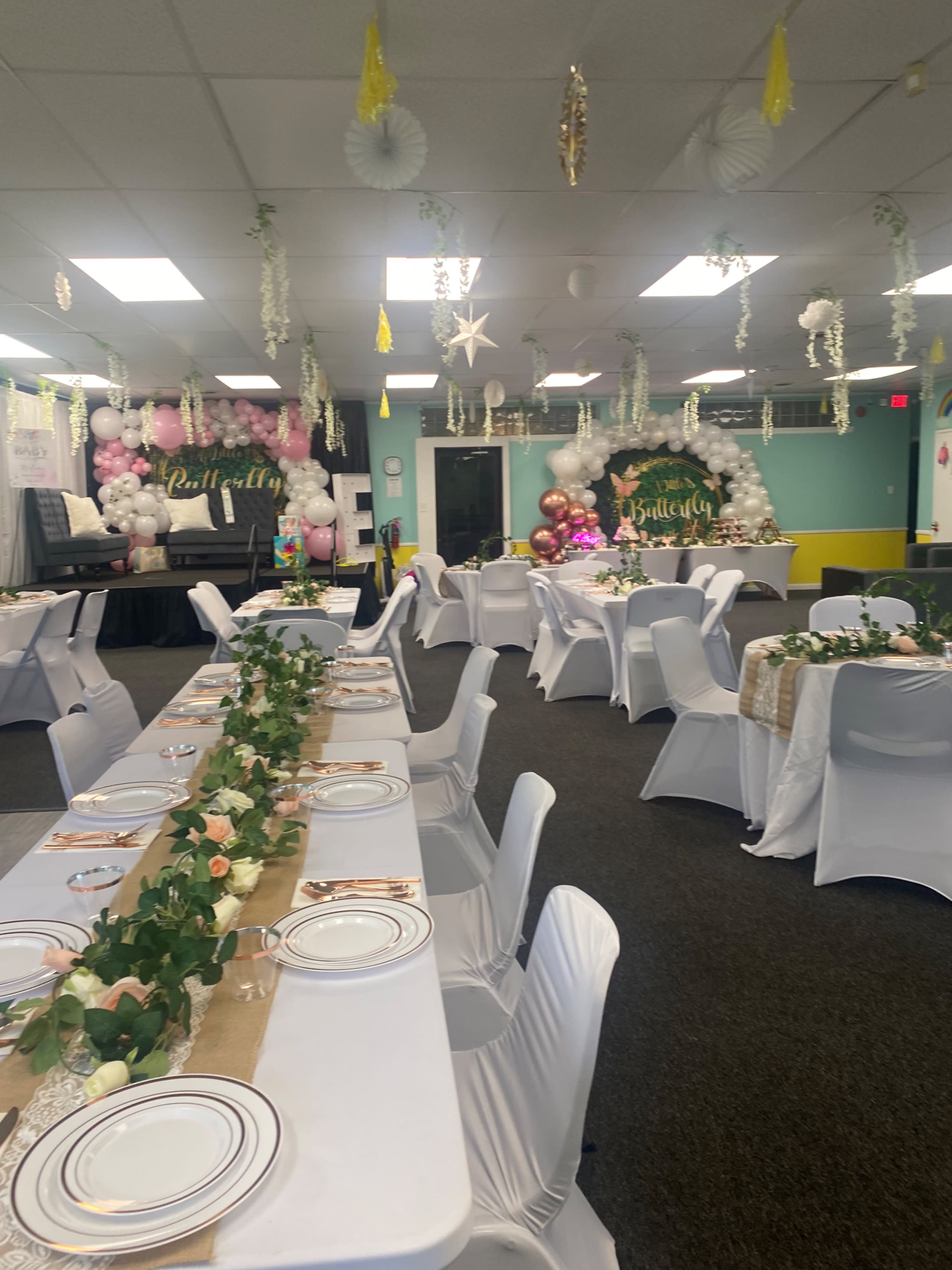 A decorated event space features a dessert table with a cake and treats, alongside a backdrop celebrating a first birthday with balloons and signage.