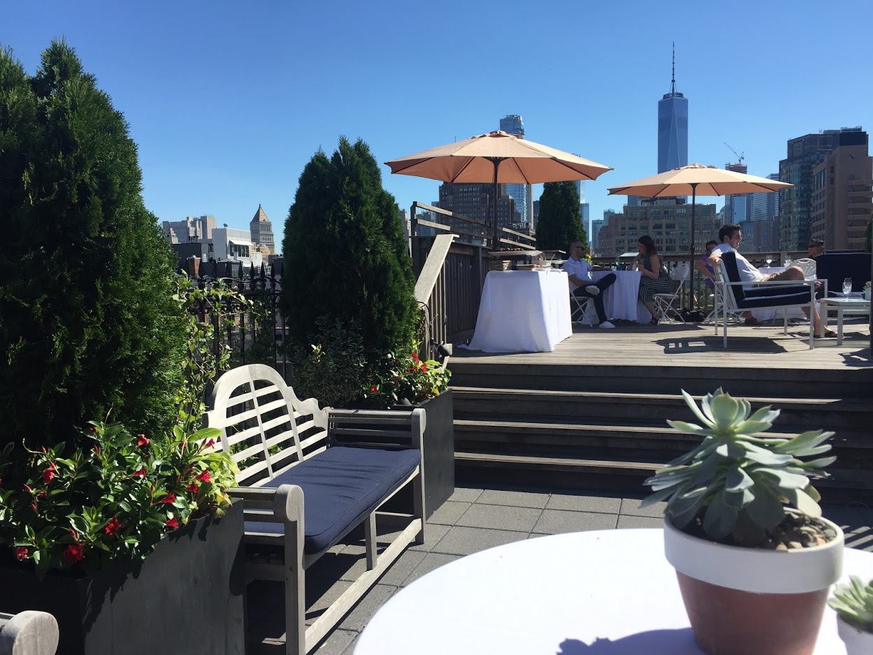 A wedding arch adorned with flowers stands on a rooftop deck, surrounded by lanterns and overlooking a city skyline under cloudy skies.