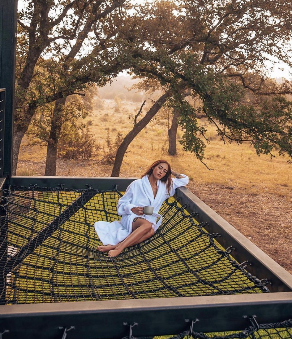 A person washes their hair while sitting in a stone bathtub outdoors, surrounded by trees and tall grass.