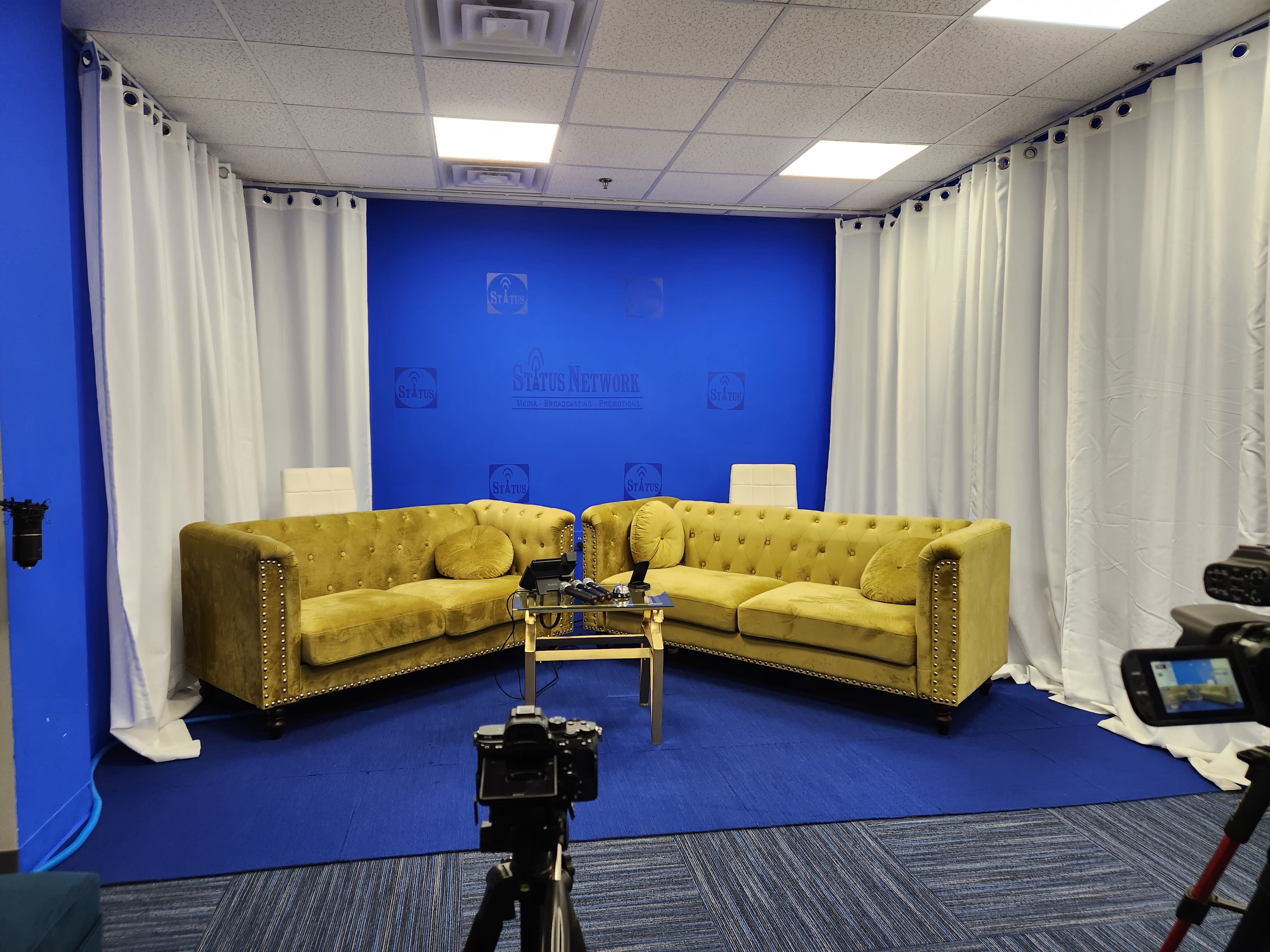 Two hosts are seated at a modern studio desk, discussing topics against a backdrop of a cityscape during a video shoot.