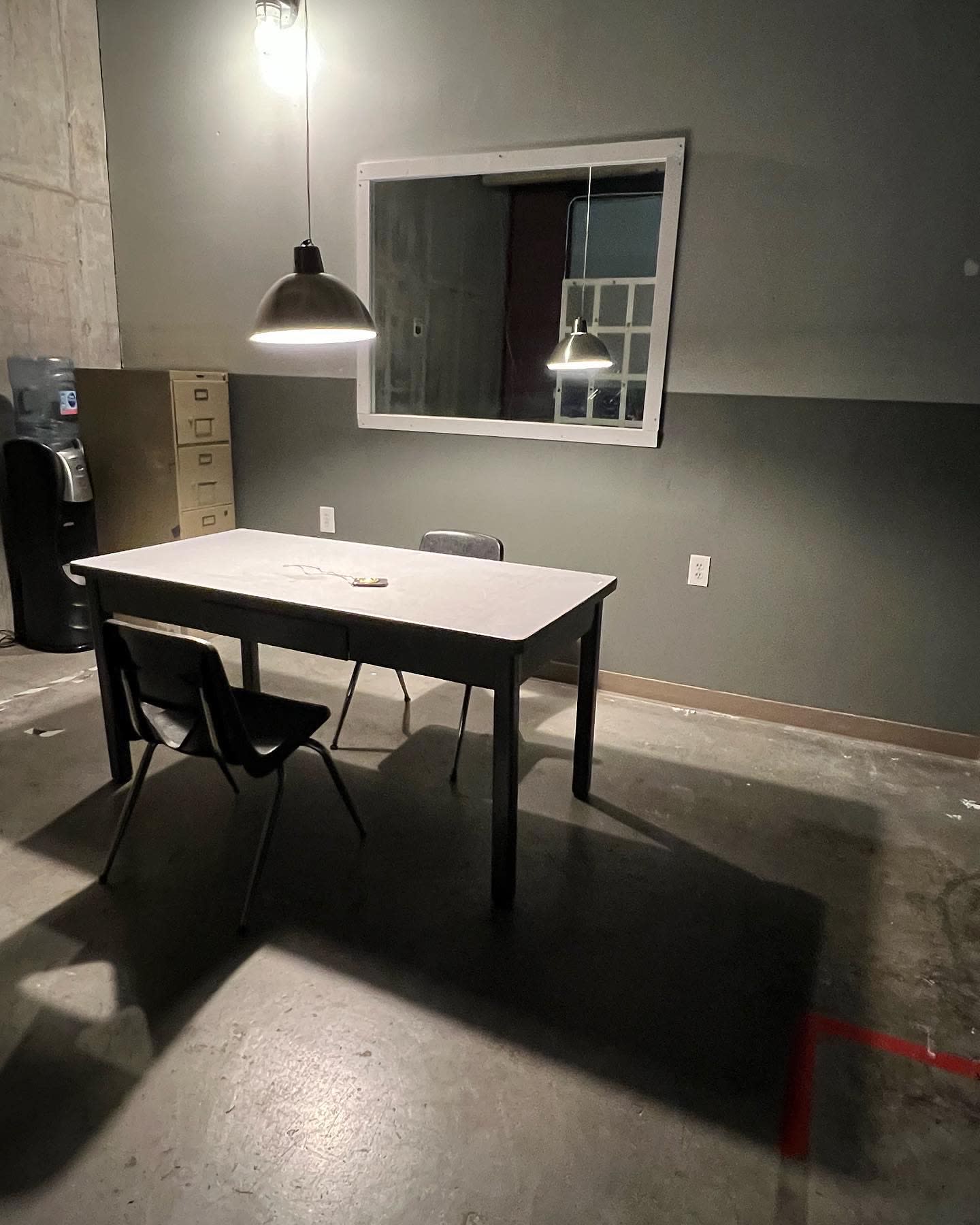 Two men sit at a table in a dimly lit interrogation room, engaged in a conversation across the table.