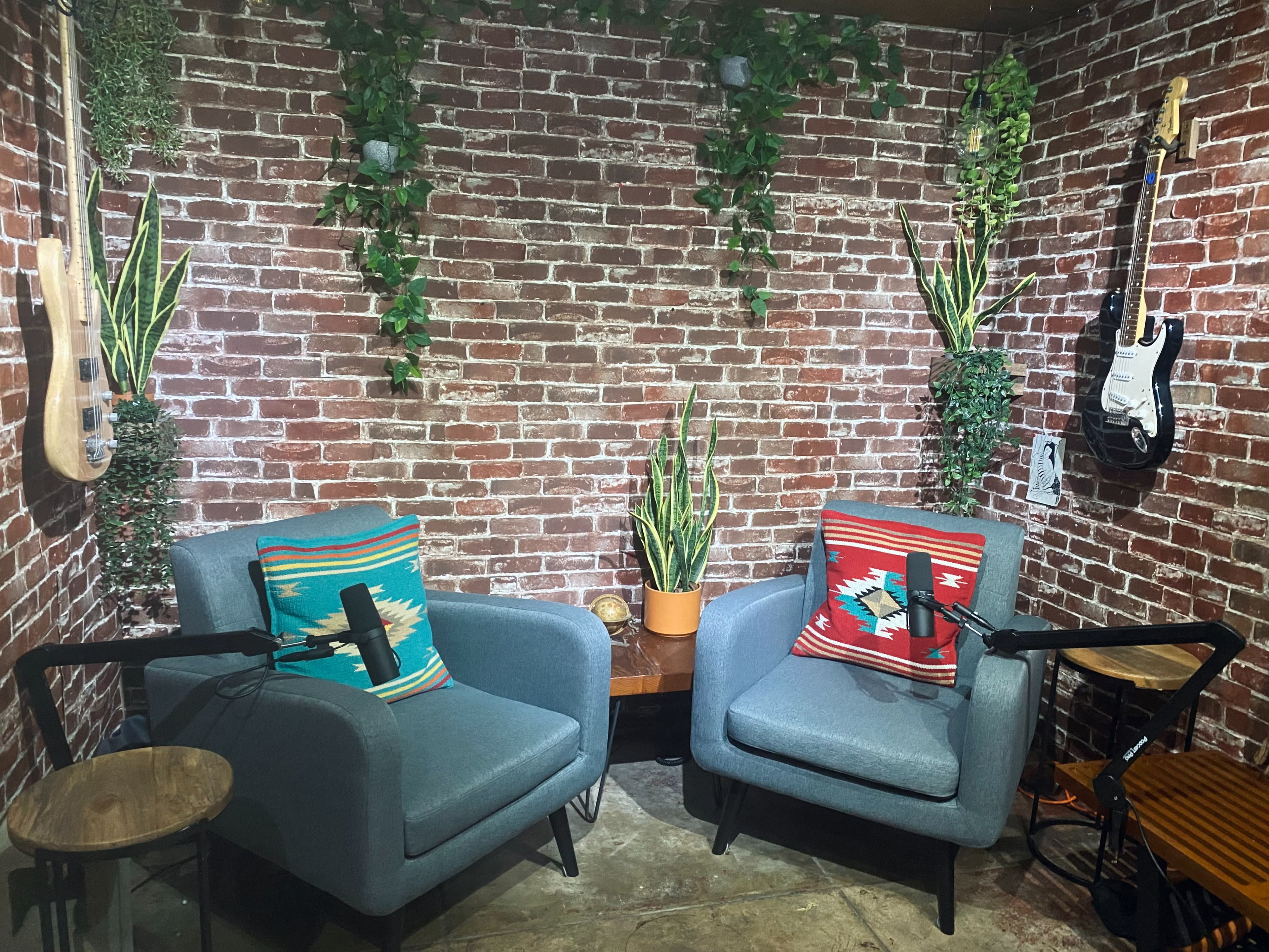Two men sit in an indoor podcast studio with a brick wall backdrop, surrounded by plants and musical instruments, ready for a recording session.