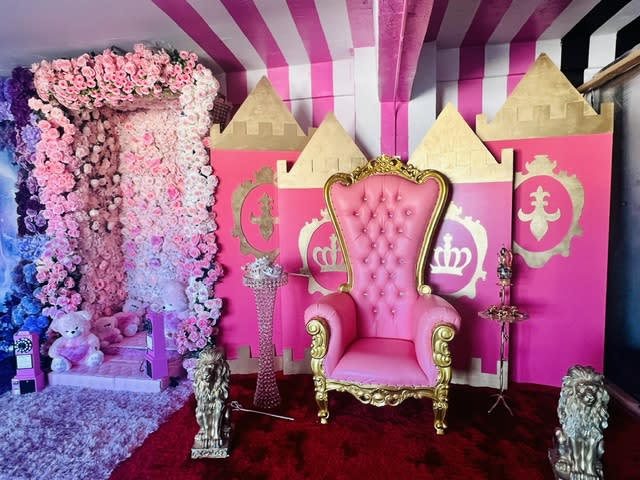 A woman sits on an ornate throne with two young girls, one holding a wand and the other clinging to her side, during a photo shoot.