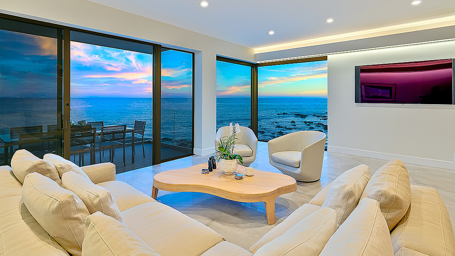 A person sits on a chair by a large window, wrapped in a dark blanket and sipping coffee while facing the ocean.