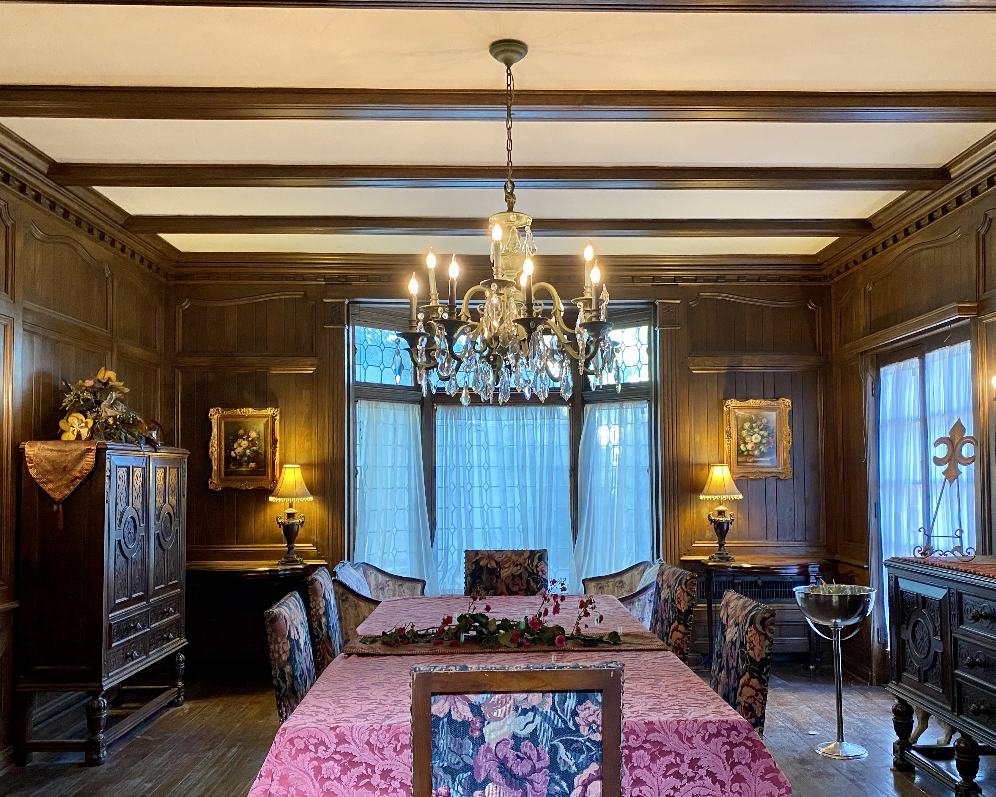 A man in a black suit writes at a wooden desk in an ornate room while surrounded by books and decorative candles.