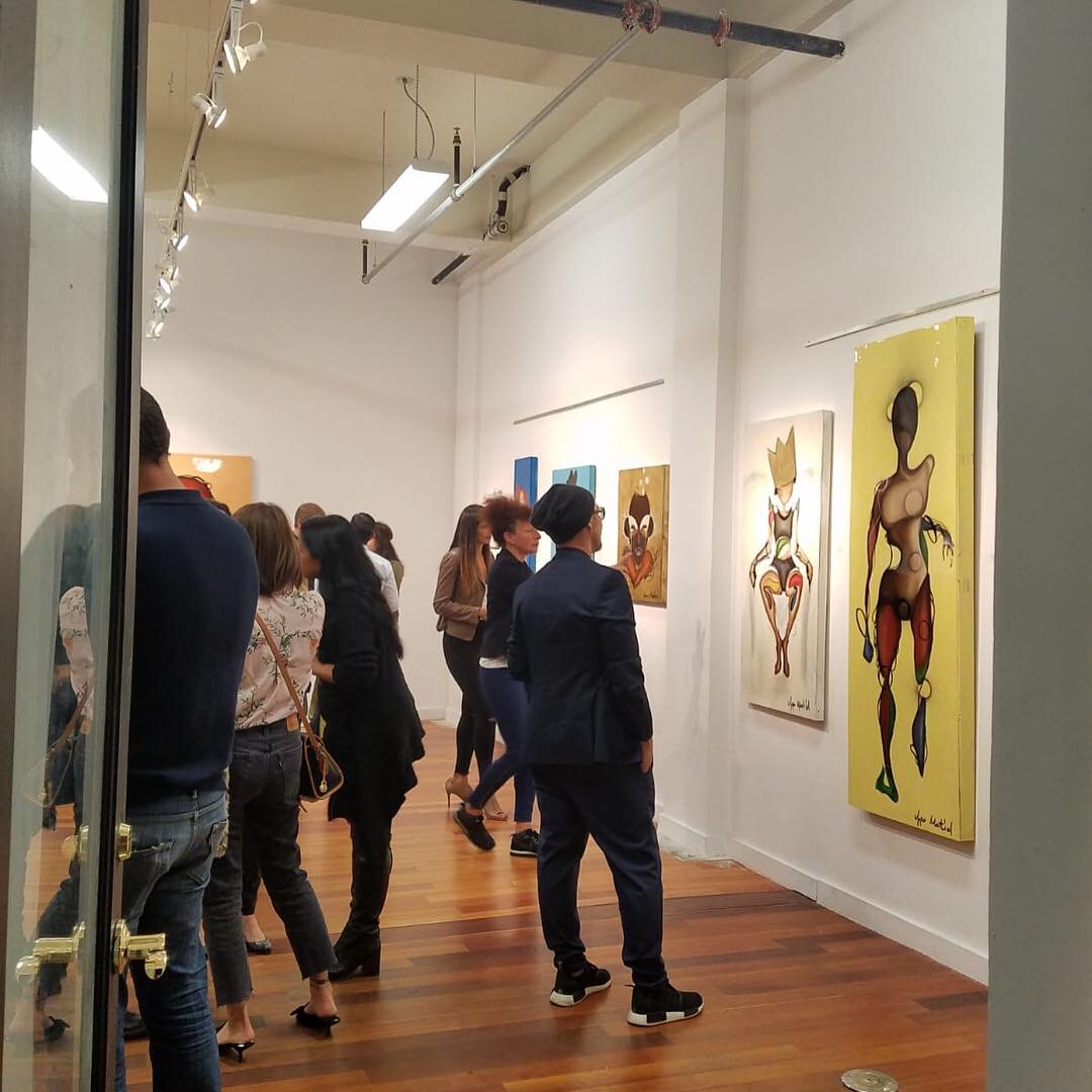 Three models showcase vibrant, traditional attire in front of striking art pieces at an exhibit.