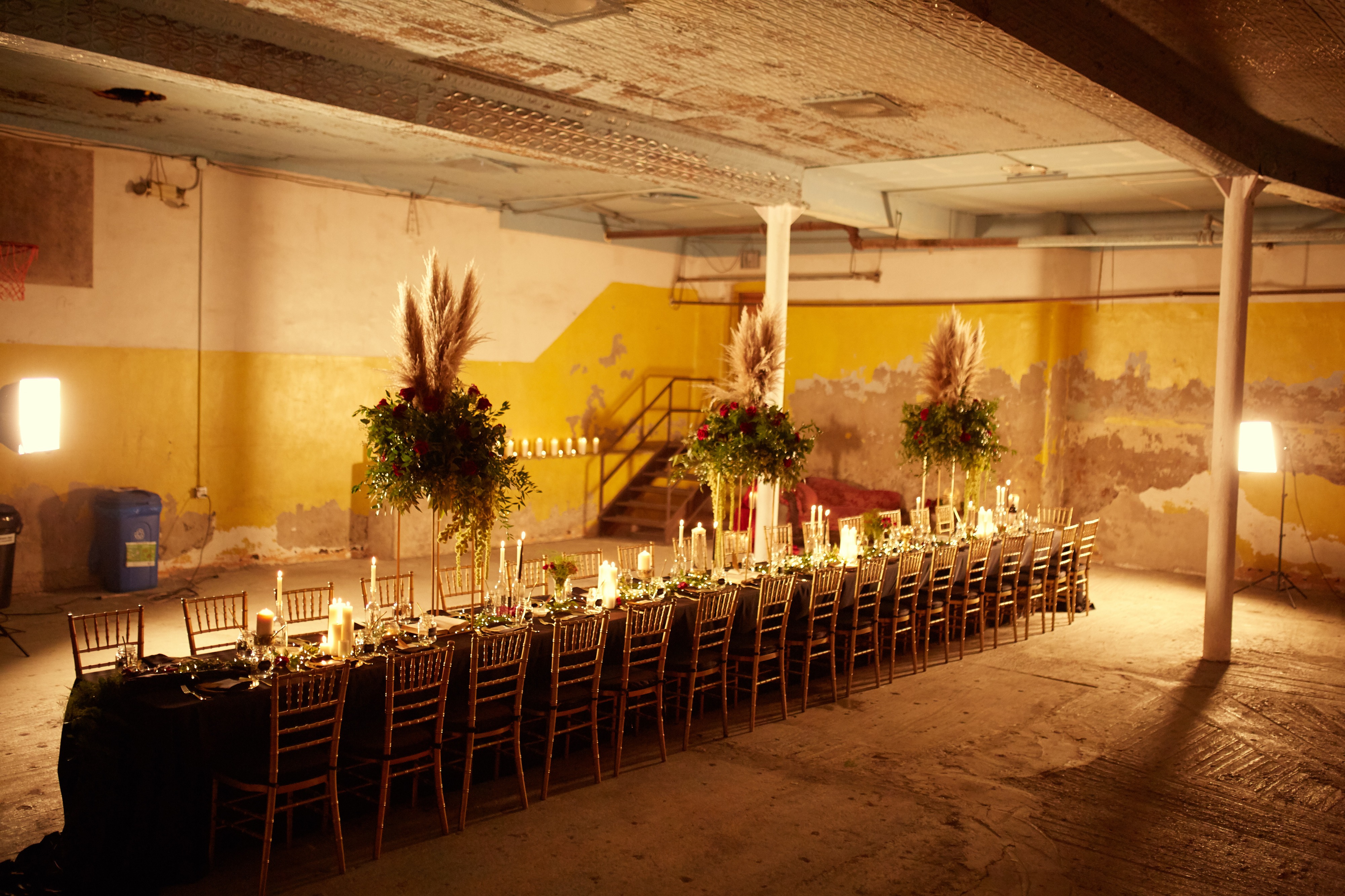 A film crew sets up a scene in a foggy warehouse, featuring two actors seated at a table while a third stands nearby.