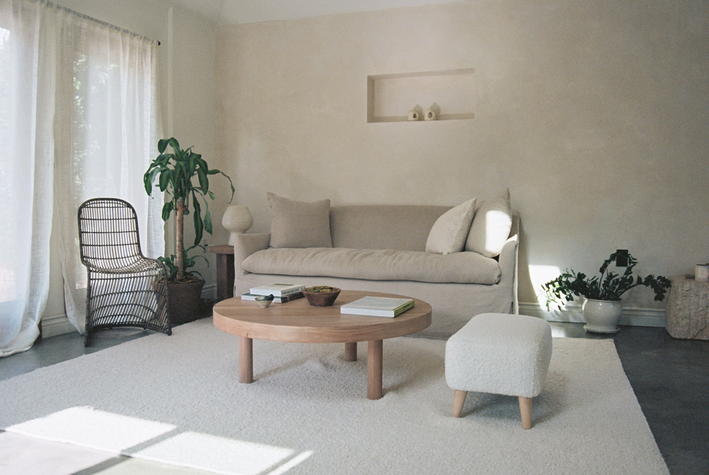 A simple bedroom features a bed with a light coverlet and a small ottoman, complemented by natural light filtering through a window.