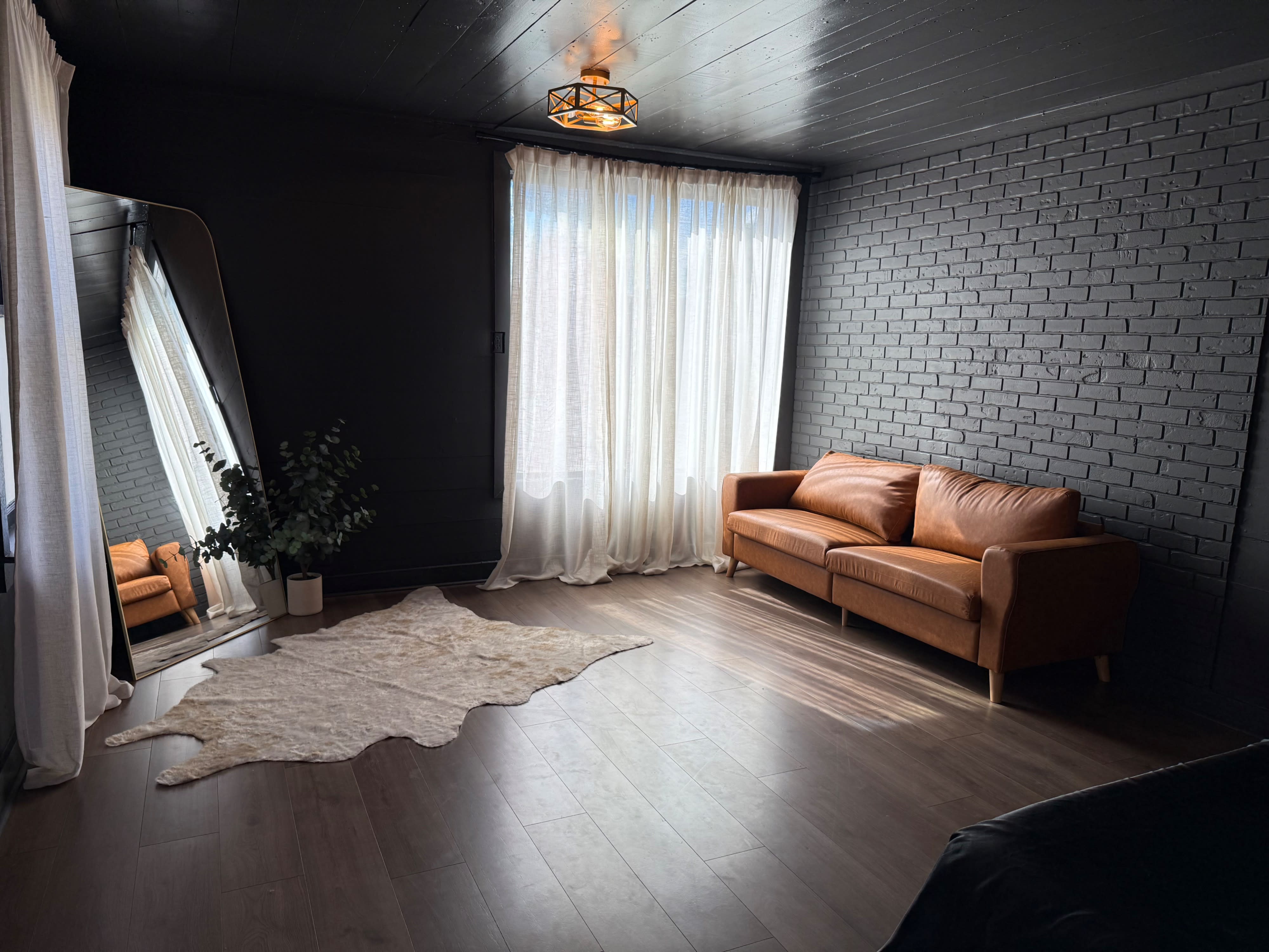 A spacious room features a brown leather sofa, a large mirror, sheer curtains, and a soft rug, creating an inviting setting for a boudoir photo shoot.