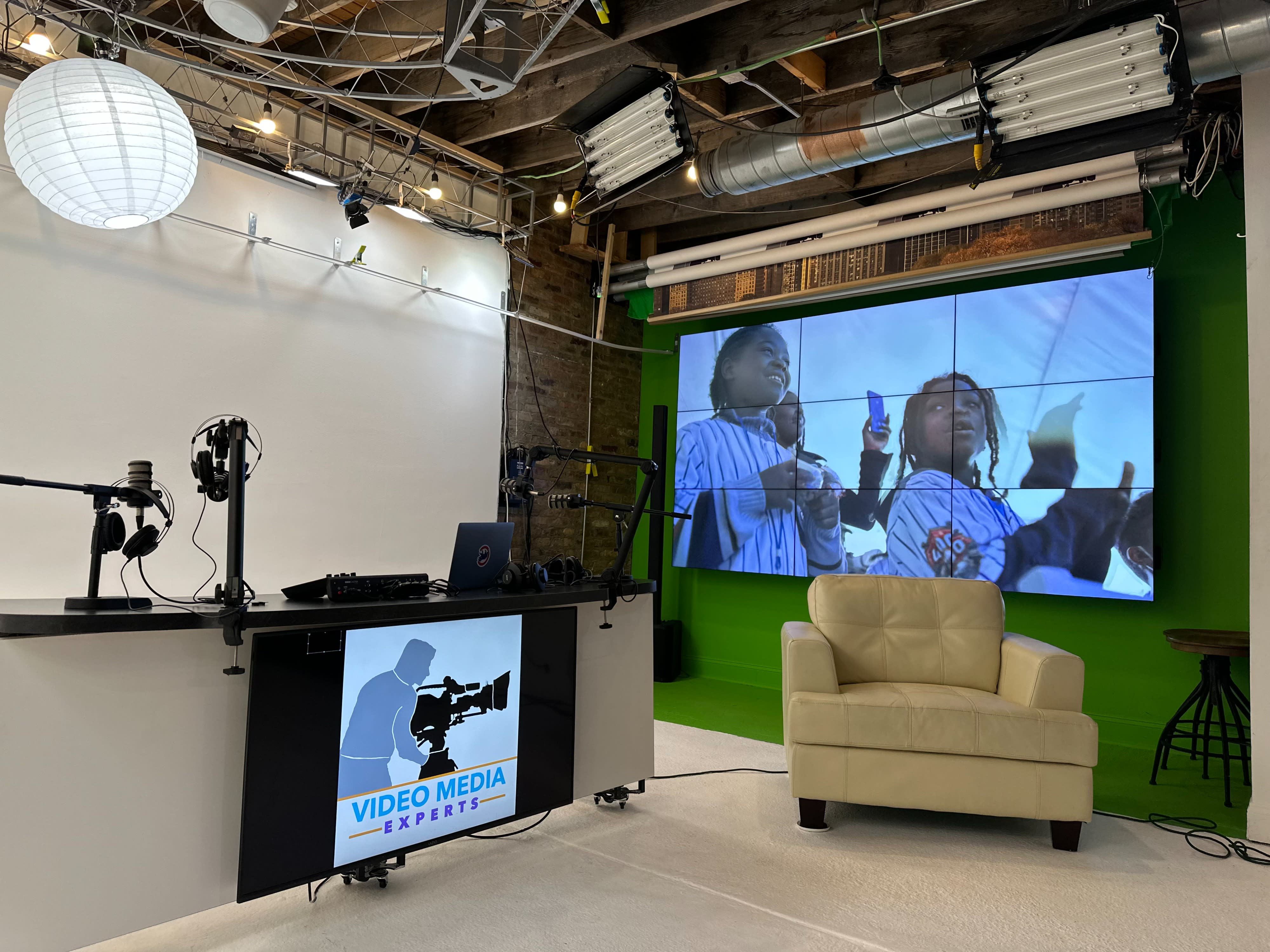 A person interviews another seated on a couch in a brightly lit studio with cameras and a green screen setup.