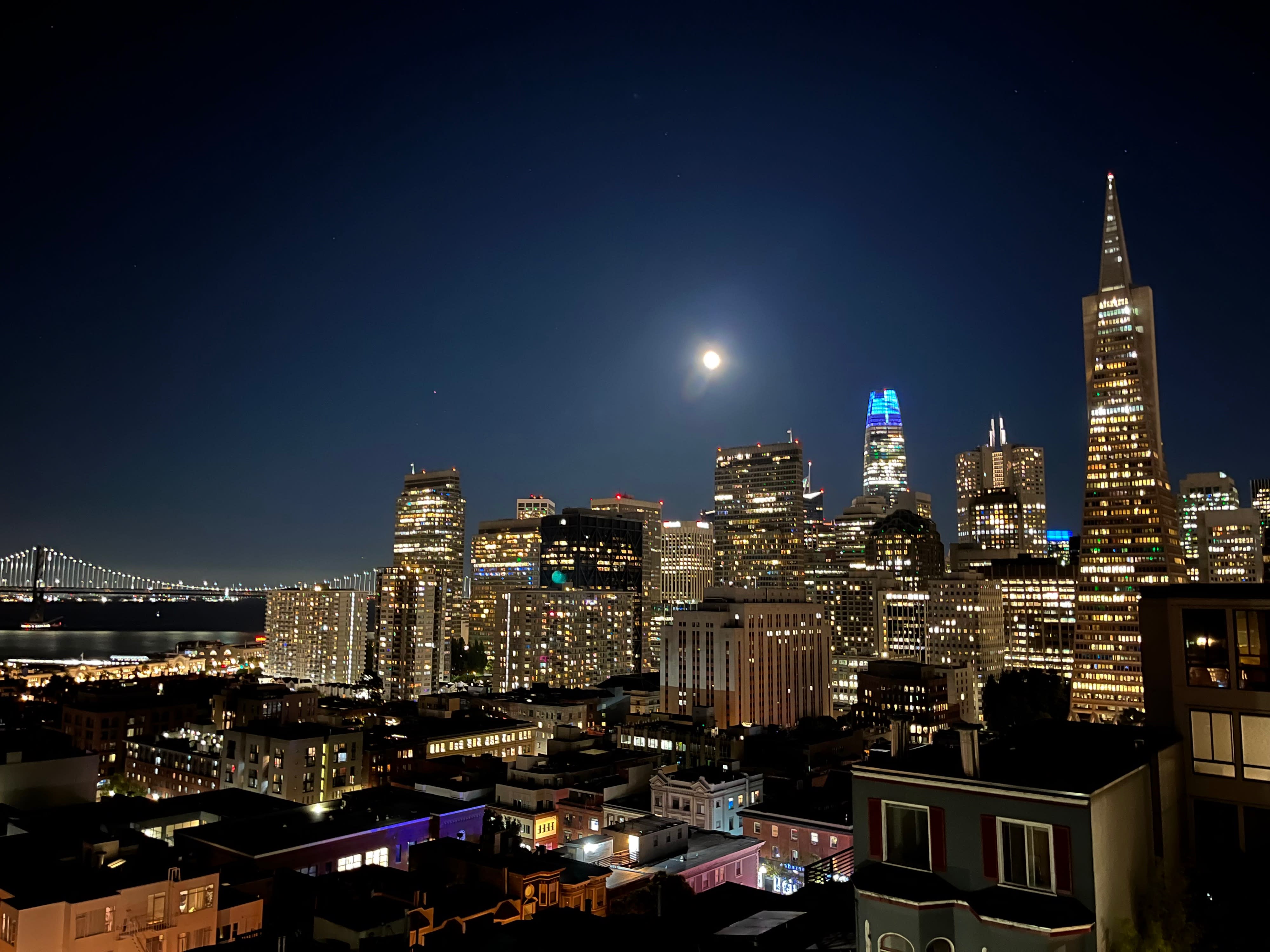 A couple stands on a rooftop deck adorned with white roses and candles, overlooking a city skyline during a wedding proposal.