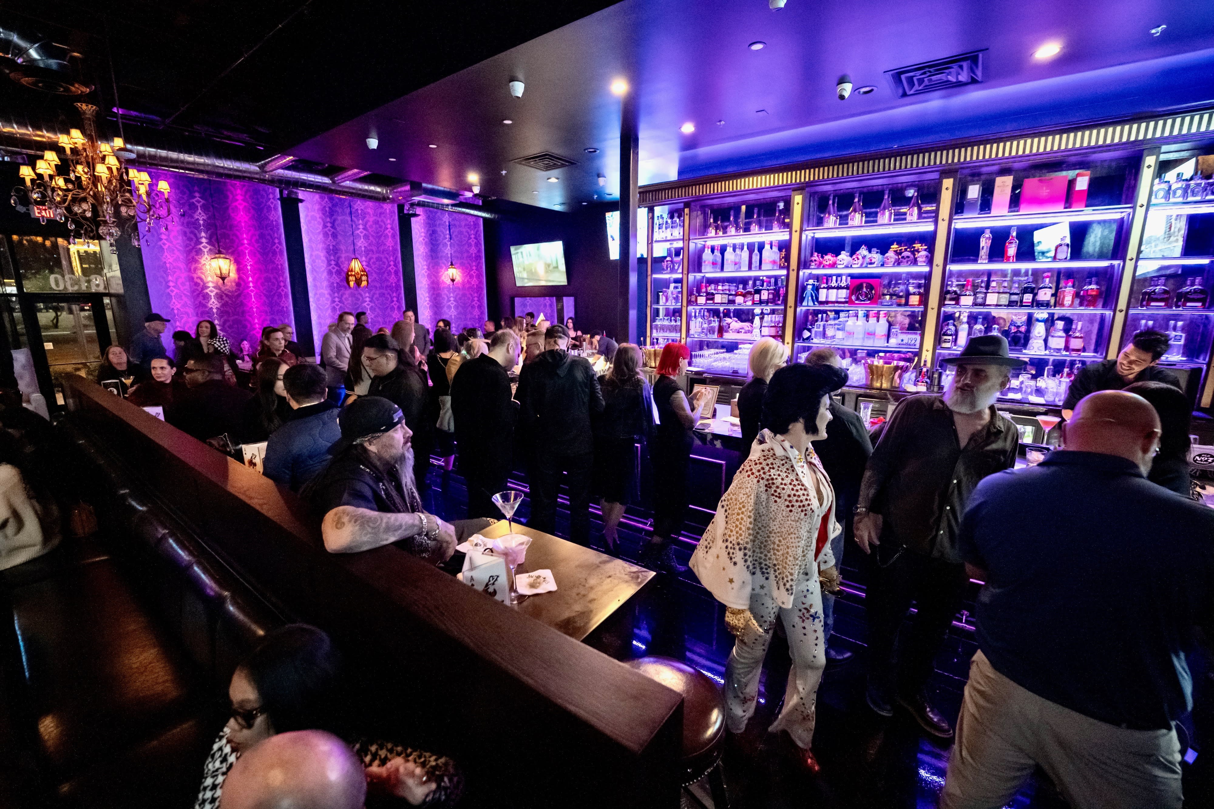 A vibrant birthday party is set up at a stylish bar adorned with purple lighting and a well-stocked backlit shelf of spirits.