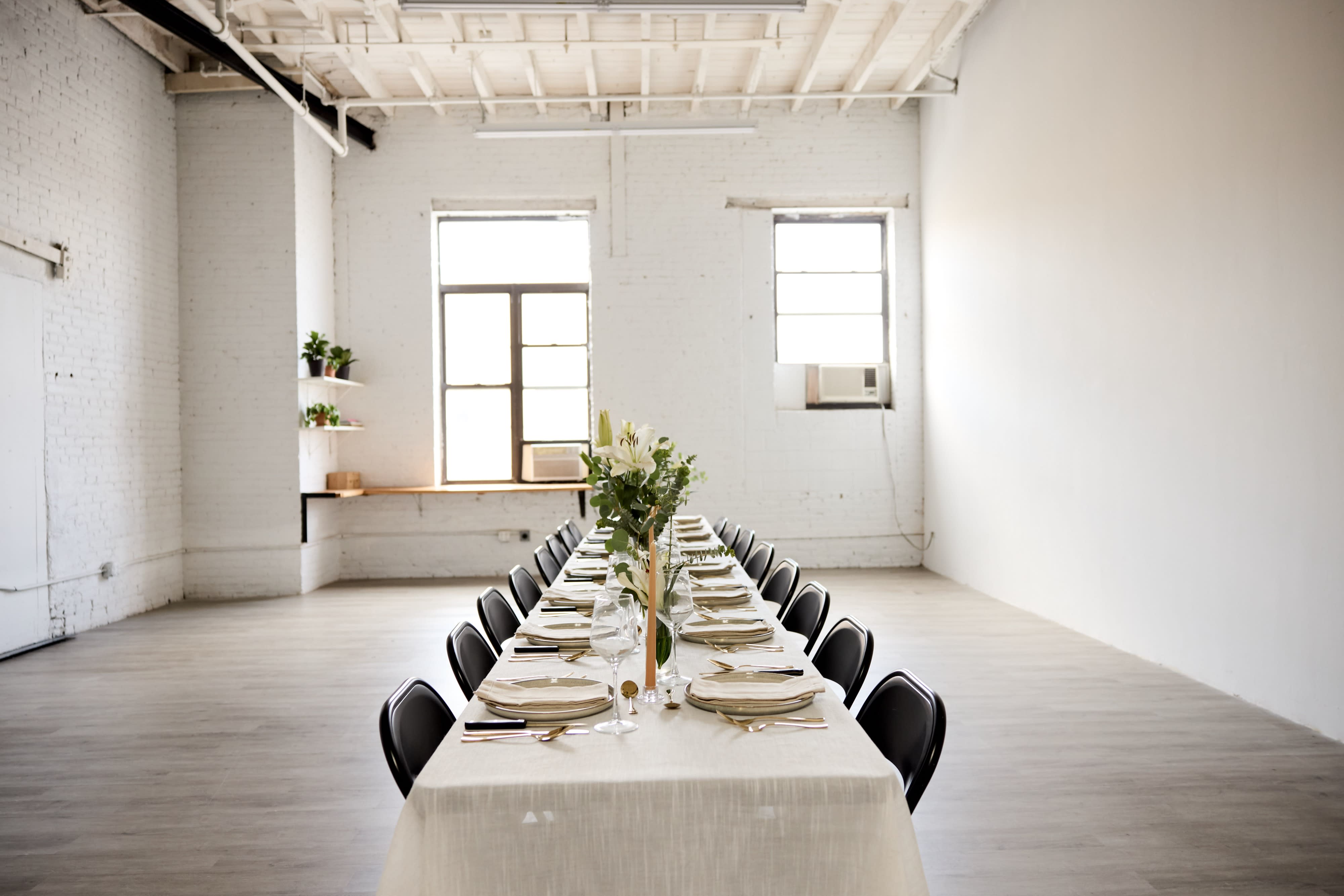 A group of people mingles around a long, elegantly set table in a well-lit, industrial-style space during a birthday dinner celebration.