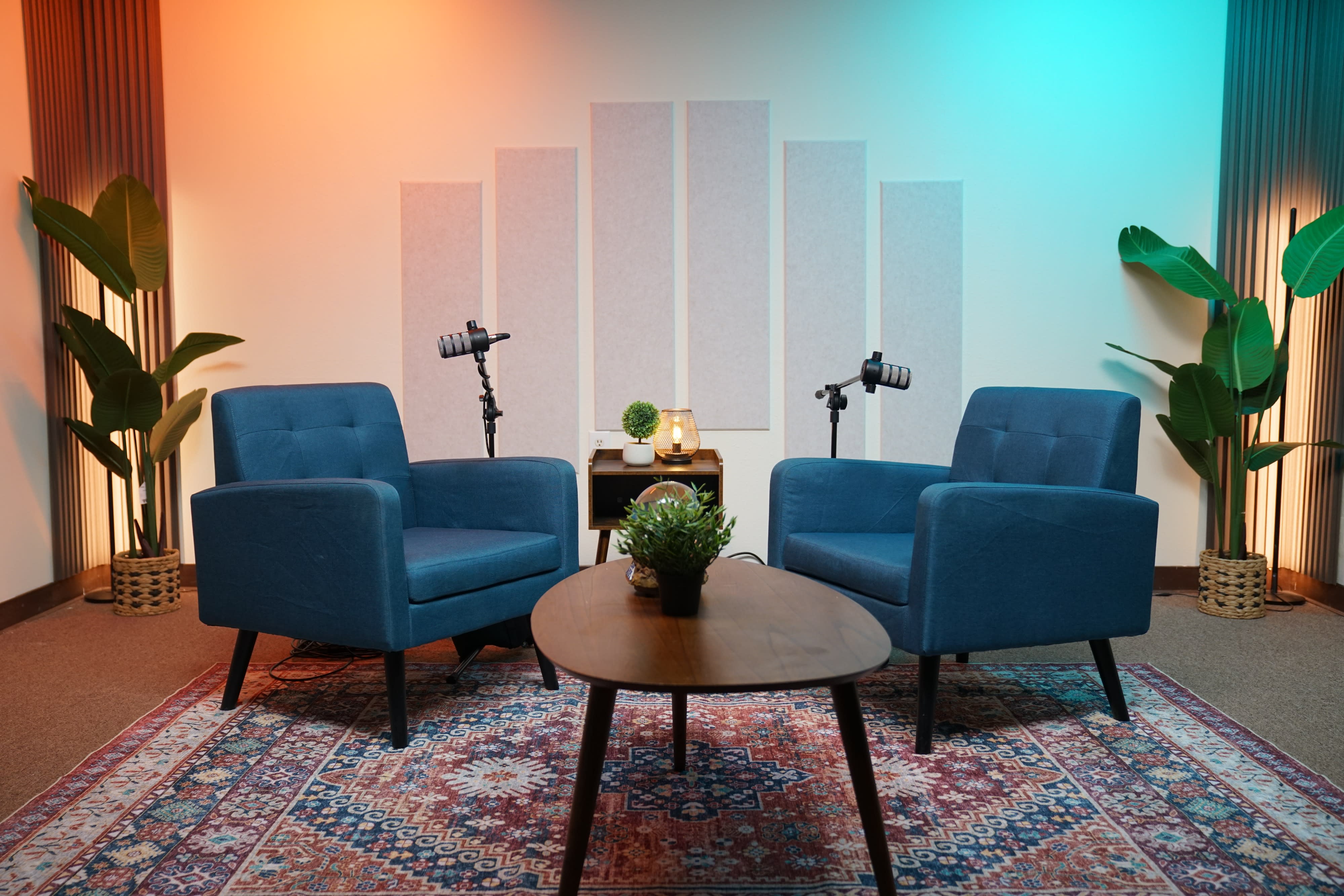 The image shows a video shoot setup featuring a cozy seating area with a blue couch, two brown chairs, and decorative plants, surrounded by lighting equipment and an engaging background.