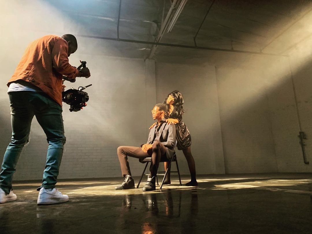A model poses confidently on a chair with one arm raised in a spacious, dimly lit environment.