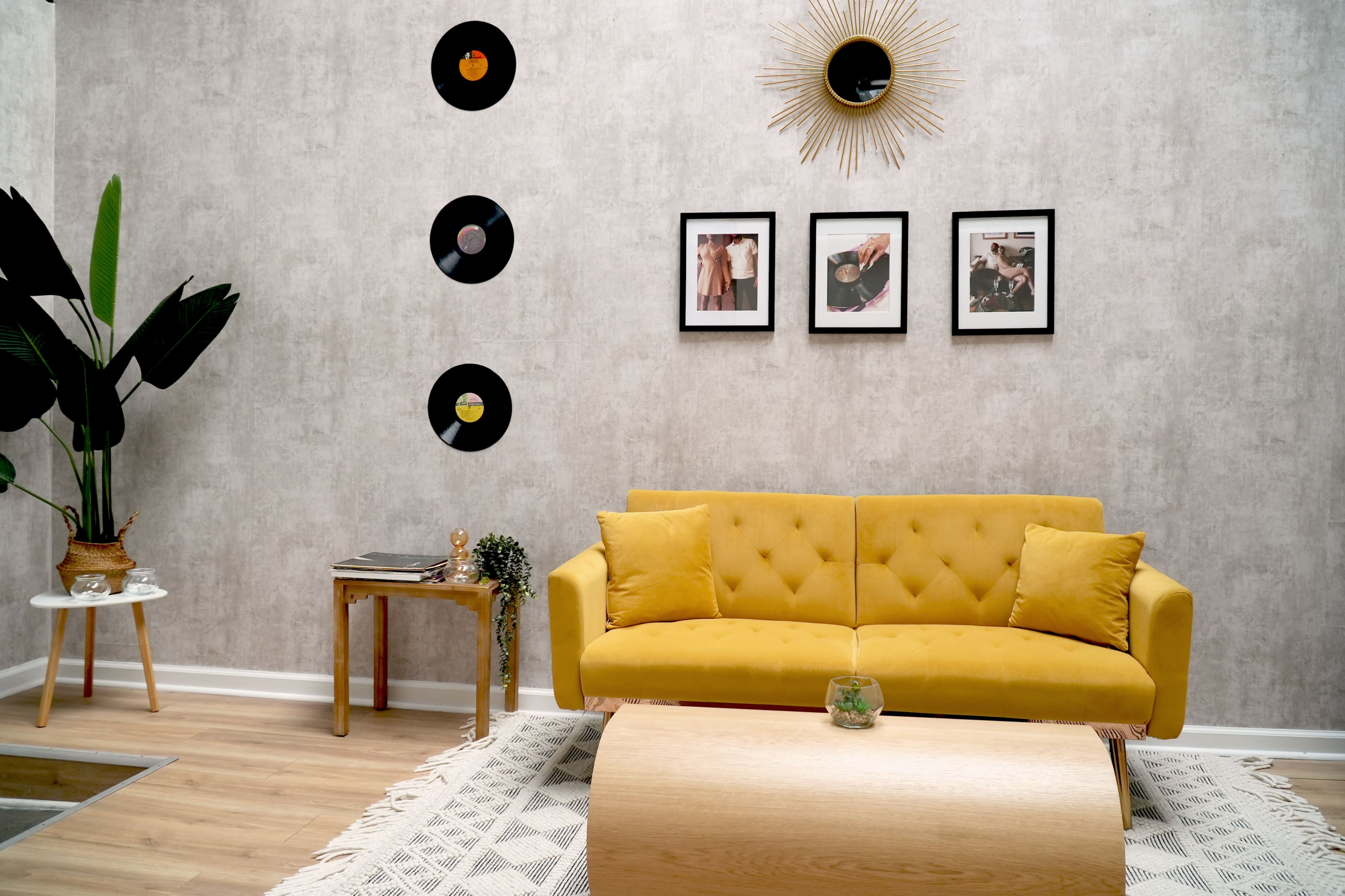 A woman sits on a yellow couch, resting her chin on her hand and looking thoughtfully at the camera in a well-lit room adorned with framed photographs.