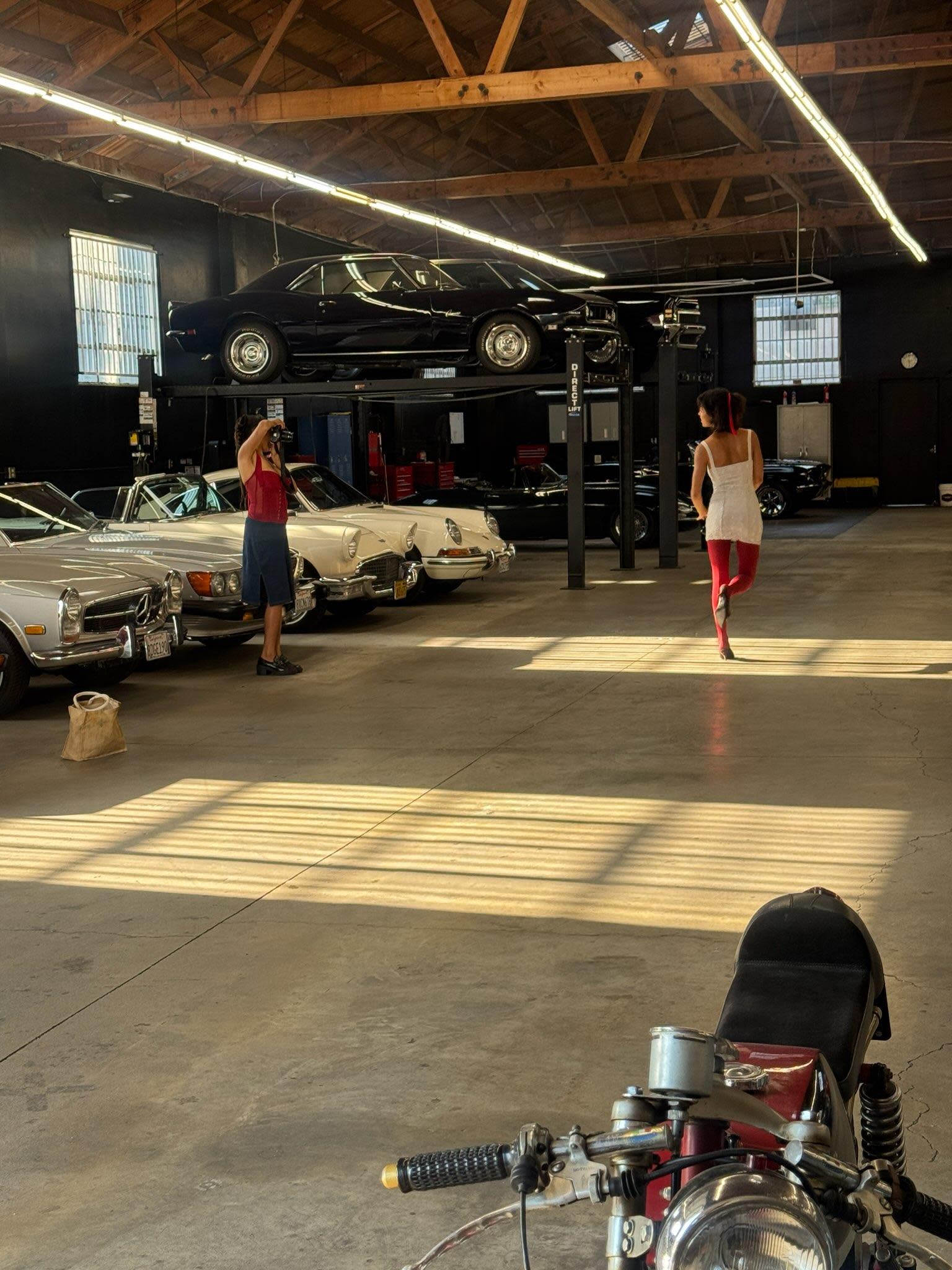 Three women hold flower bouquets while posing together at a launch event inside a spacious venue filled with vintage cars.