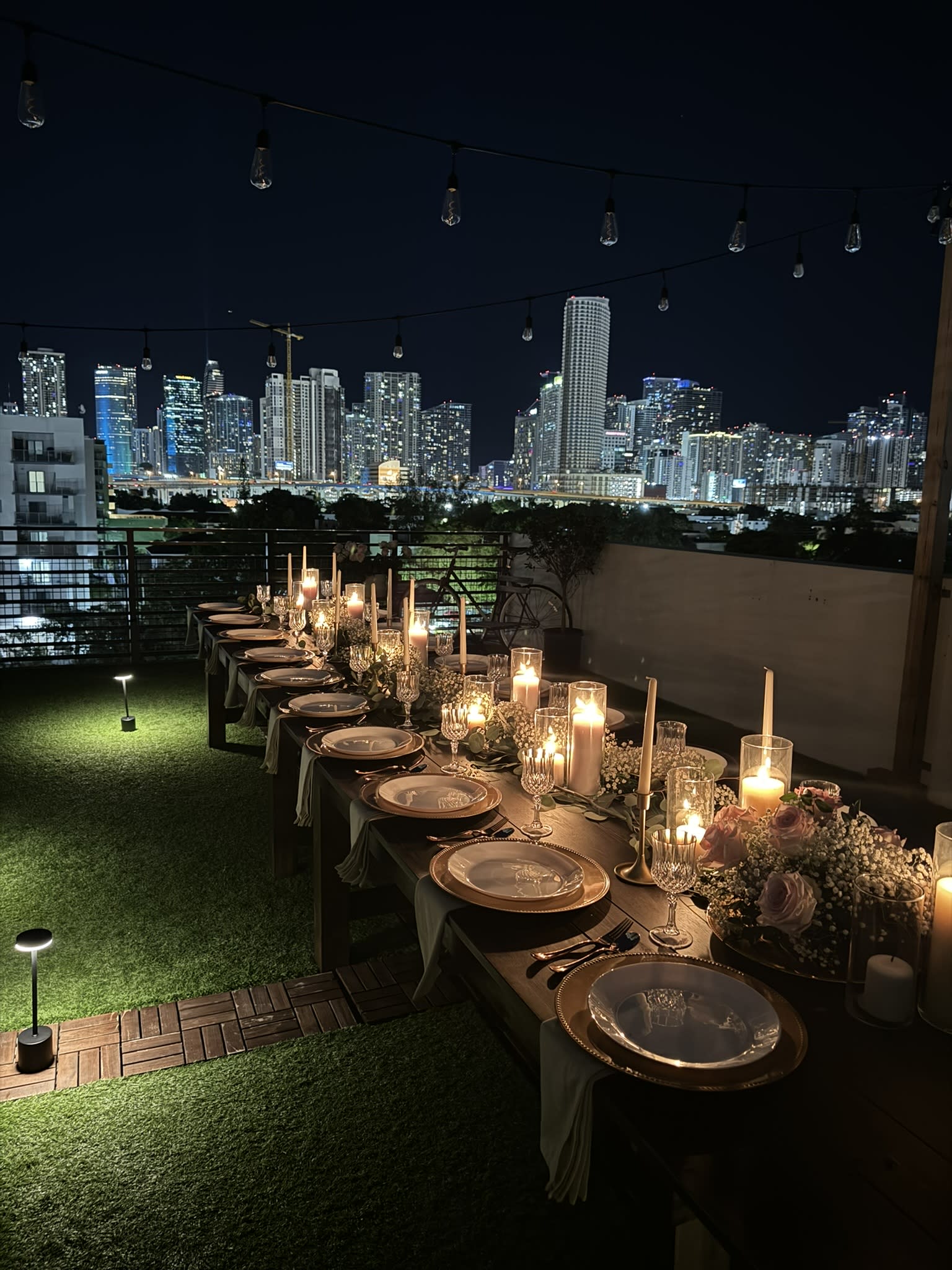 A woman stands next to a beautifully arranged table on a rooftop, decorated for a birthday party with flowers and candles, overlooking a city skyline at night.