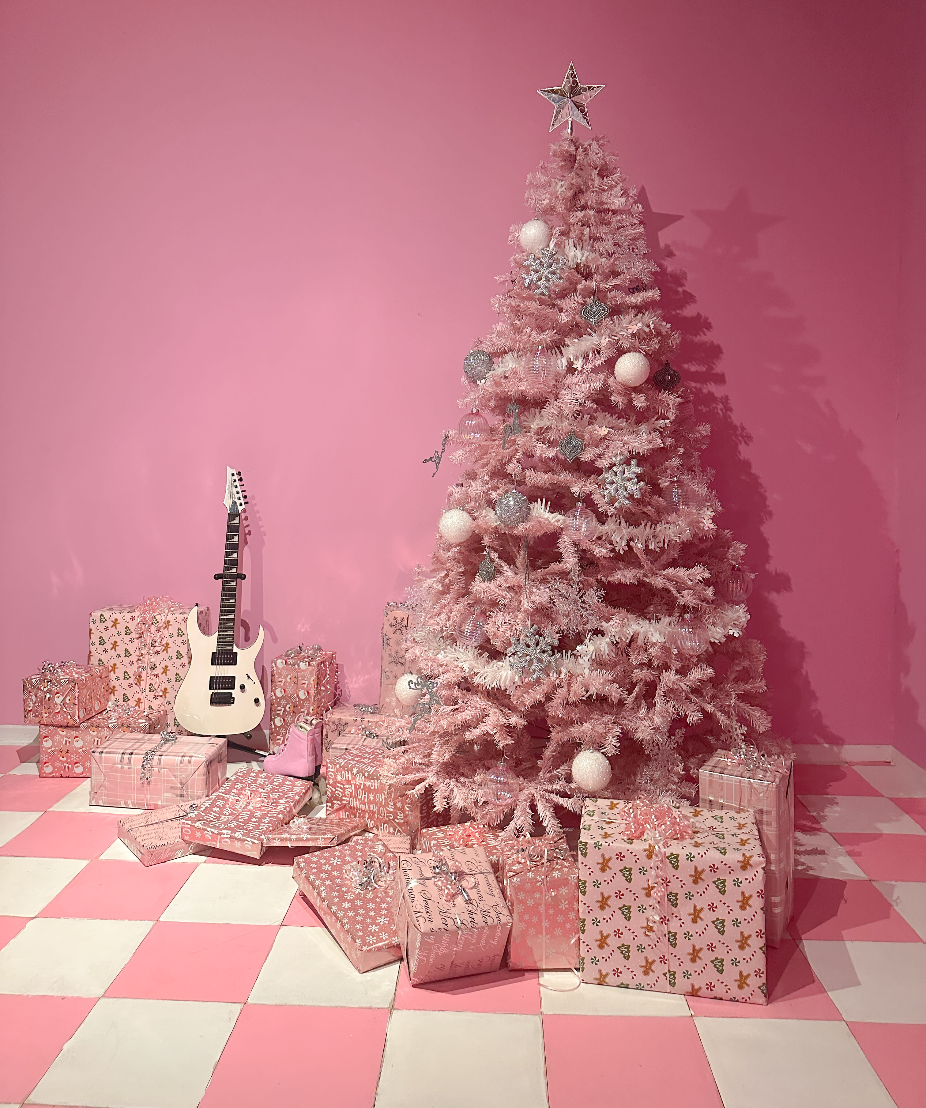 A model poses next to a pink Christmas tree while decorating it with ornaments in a pink-themed studio.
