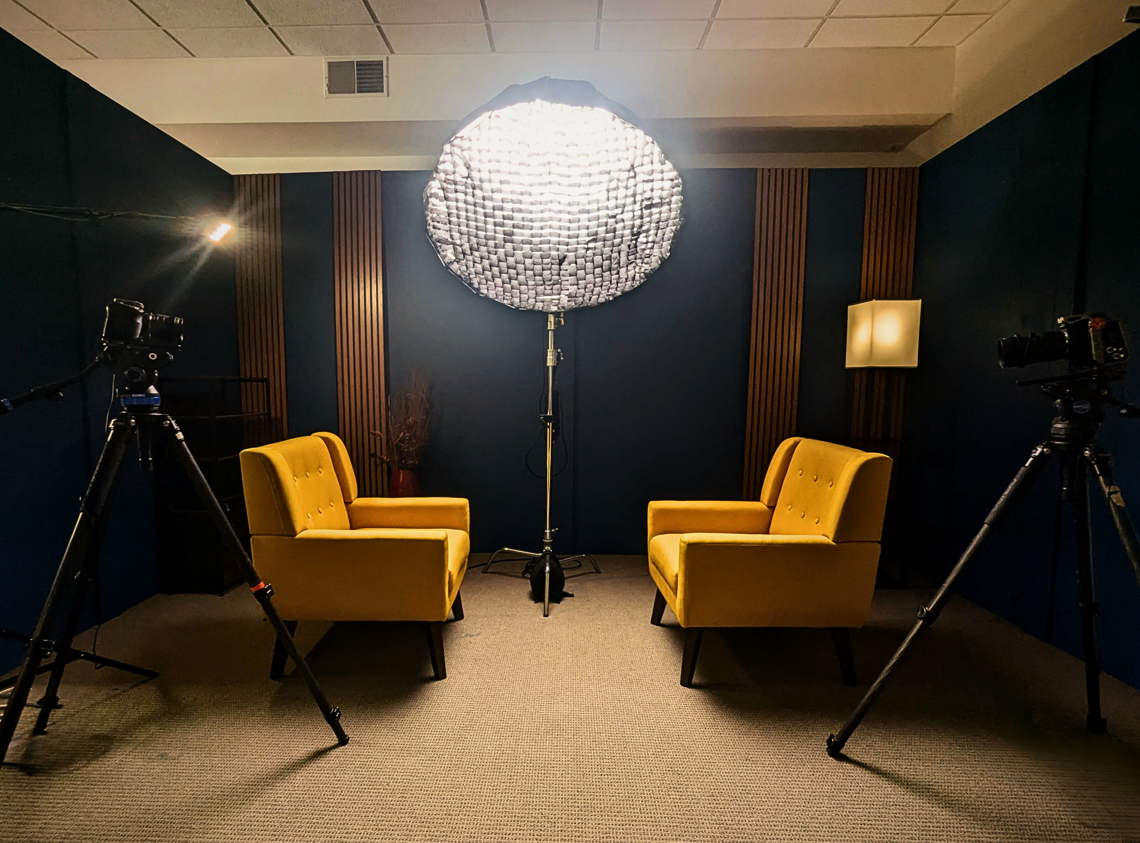 A video shoot setup features a bright yellow chair in focus, surrounded by professional lighting, cameras, and a microphone.