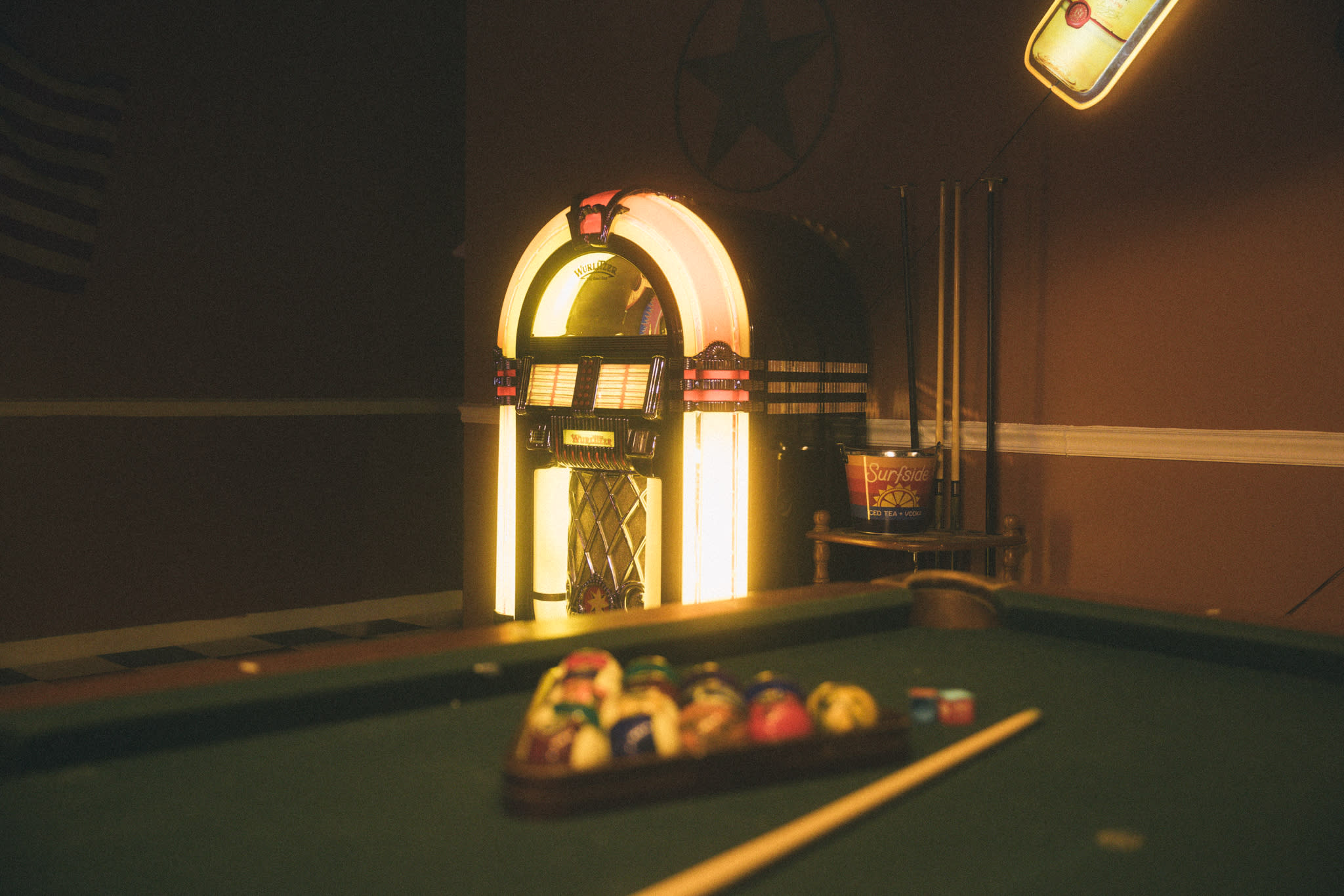 A woman in casual attire poses with a cue stick near a dartboard and disco balls in a retro bar setting.