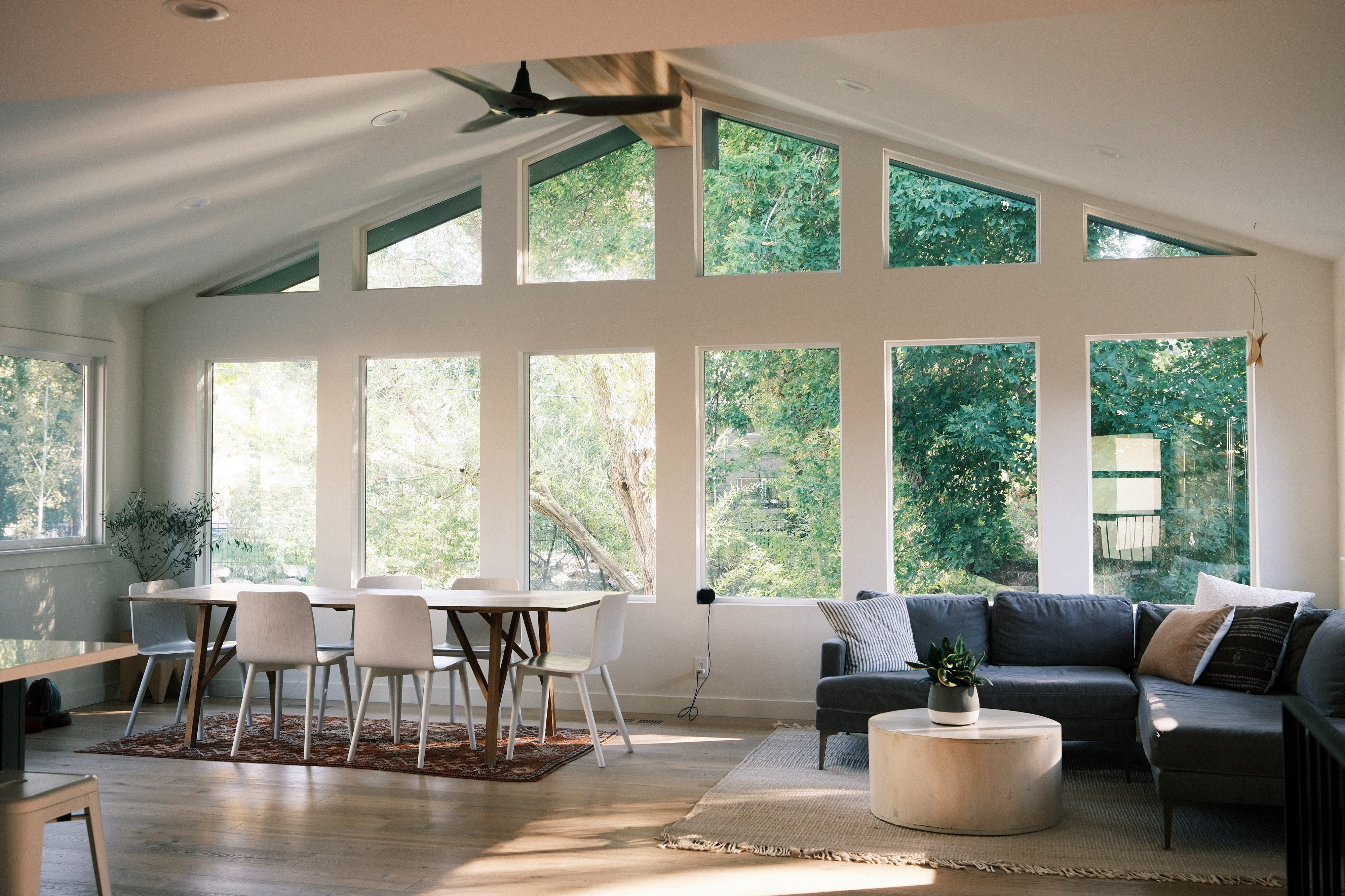 A well-lit living space features a dining table with chairs, a cozy seating area, and large windows overlooking greenery.