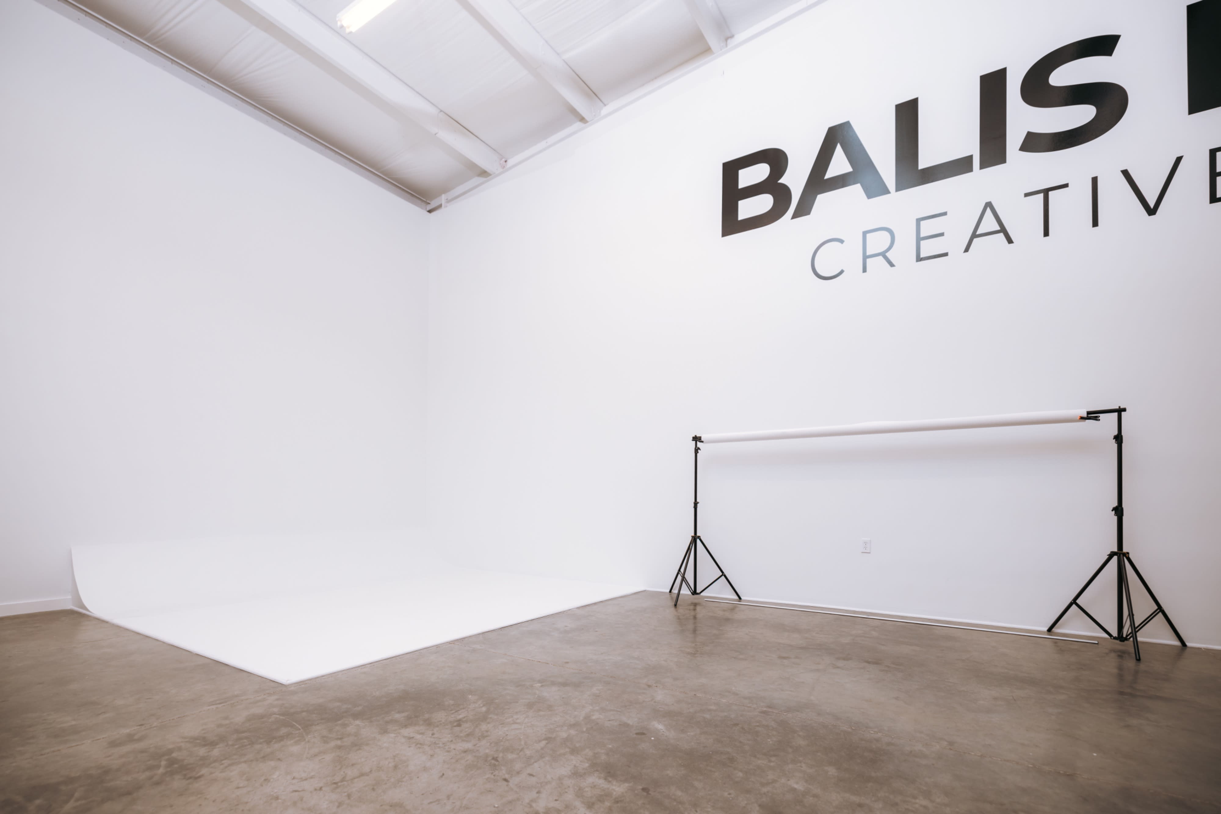The image shows a video shoot setup in a studio marked "BALS HOUSE CREATIVE STUDIO," featuring a white backdrop, a light stand, and props including pink dumbbells.