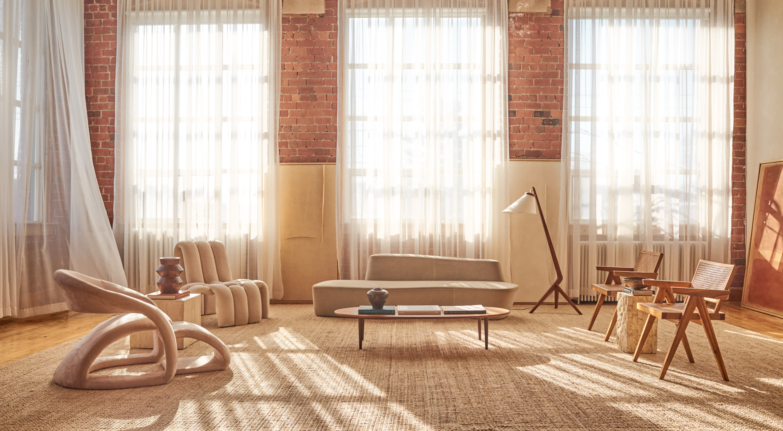 A person poses on a curved sofa in a bright room with large windows and soft curtains.