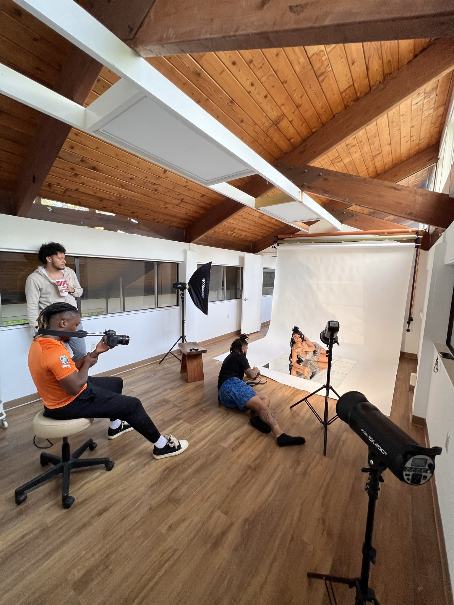 Two women are posing for a photoshoot, one sitting elegantly in a chair while the other kneels beside her on the floor.
